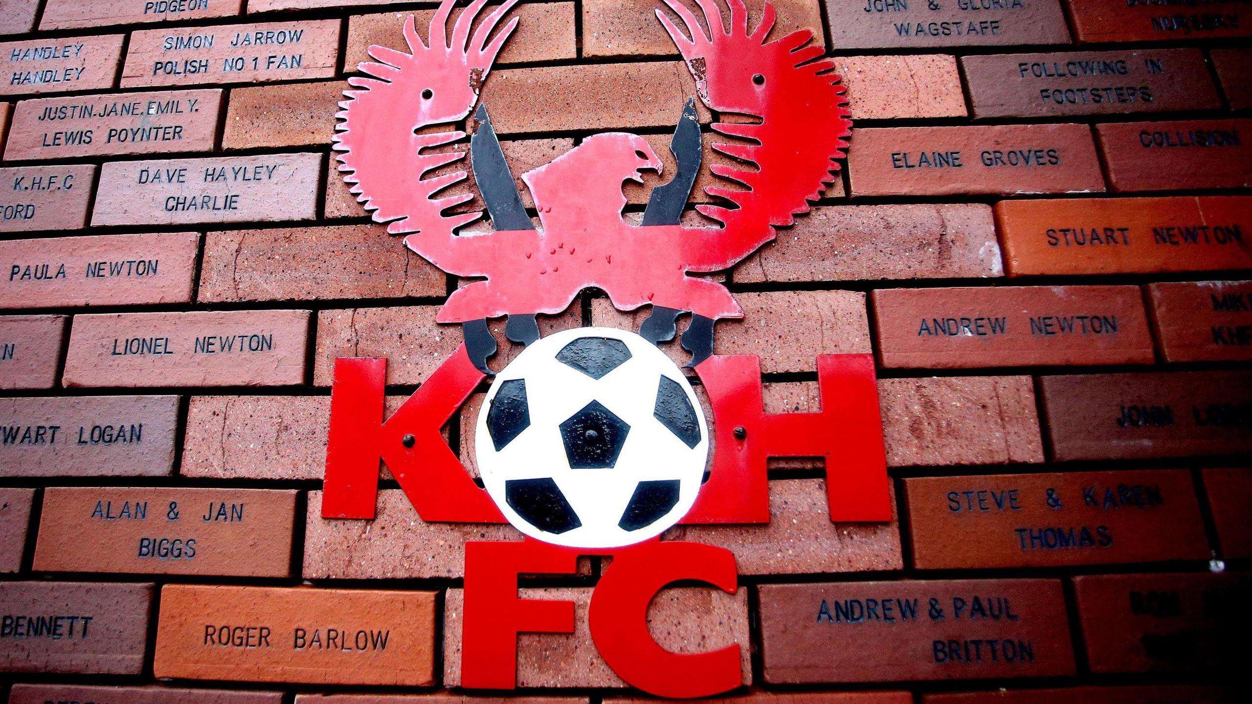 Badge of Kidderminster Harriers with a bird on top of a black and white football with the letters K and H either side of it and FC underneath. The badge is attached to a wall of bricks showing names of supporters.