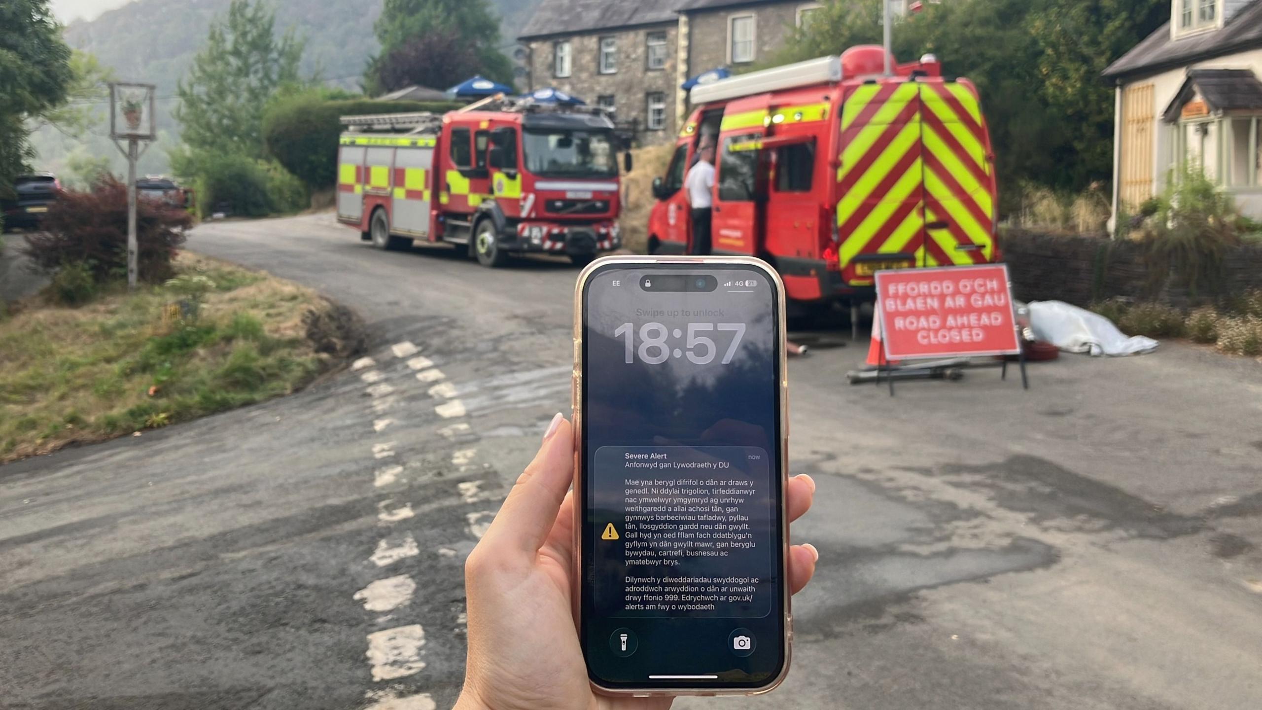 A phone screen with an emergency alert on is seen on a phone, with fire engines behind