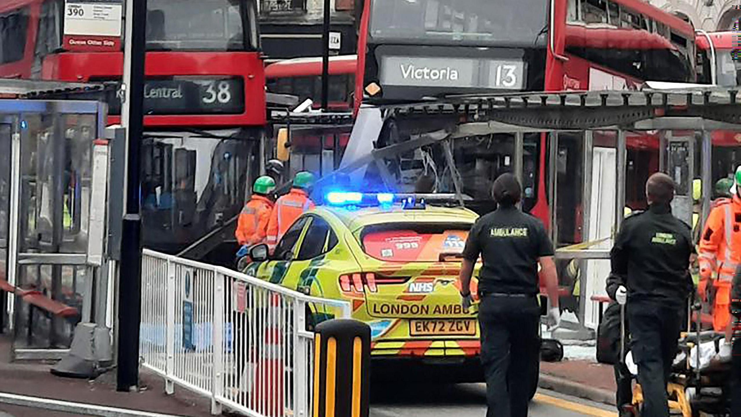 The scene of a bus crash at London Victoria Bus Station on 29 January. Paramedics are in the foreground. A London Ambulance car is parked in front of a red double decker bus that has crashed into a glass and metal bus shelter. Other emergency workers in orange high visibility clothes are at the scene.