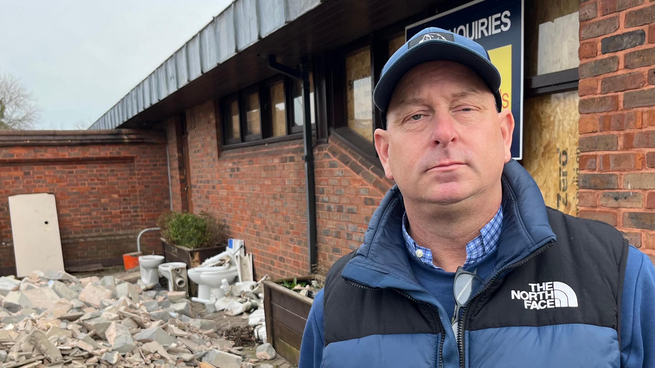 Andy Simmonds, 45, wearing a blue baseball cap, blue body warmer over a blue top with a blue and white chequered shirt underneath. He is standing in front of a corner of the old brick pub with a large pile of debris and rubble to the left, including white porcelain toilets and bricks.