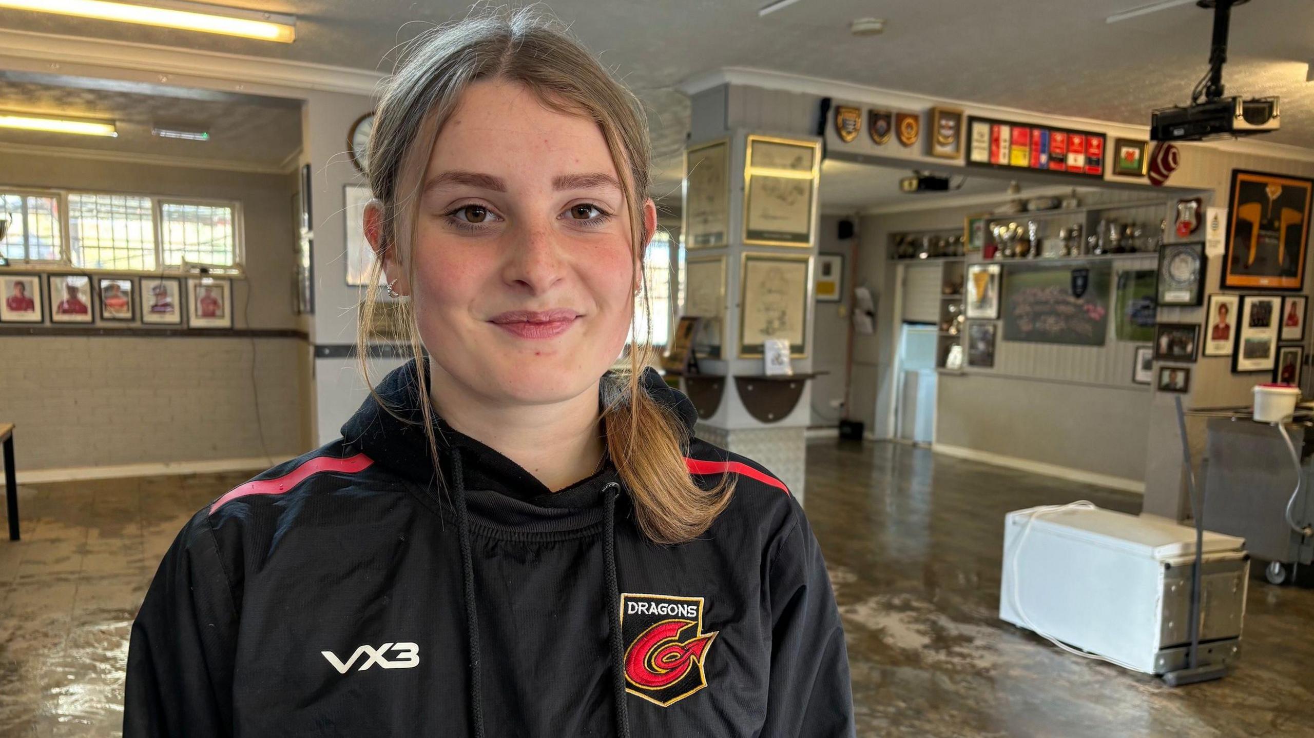 A young lady with blonde hair tied back in a ponytail is looking at the camera. She's wearing a black hoodie with the Dragons rugby team logo on it. In the background is room that has been affected by floodwater. There's a white fridge on the floor to the right of frame and there are many different types of photo frames and shields on the walls. 