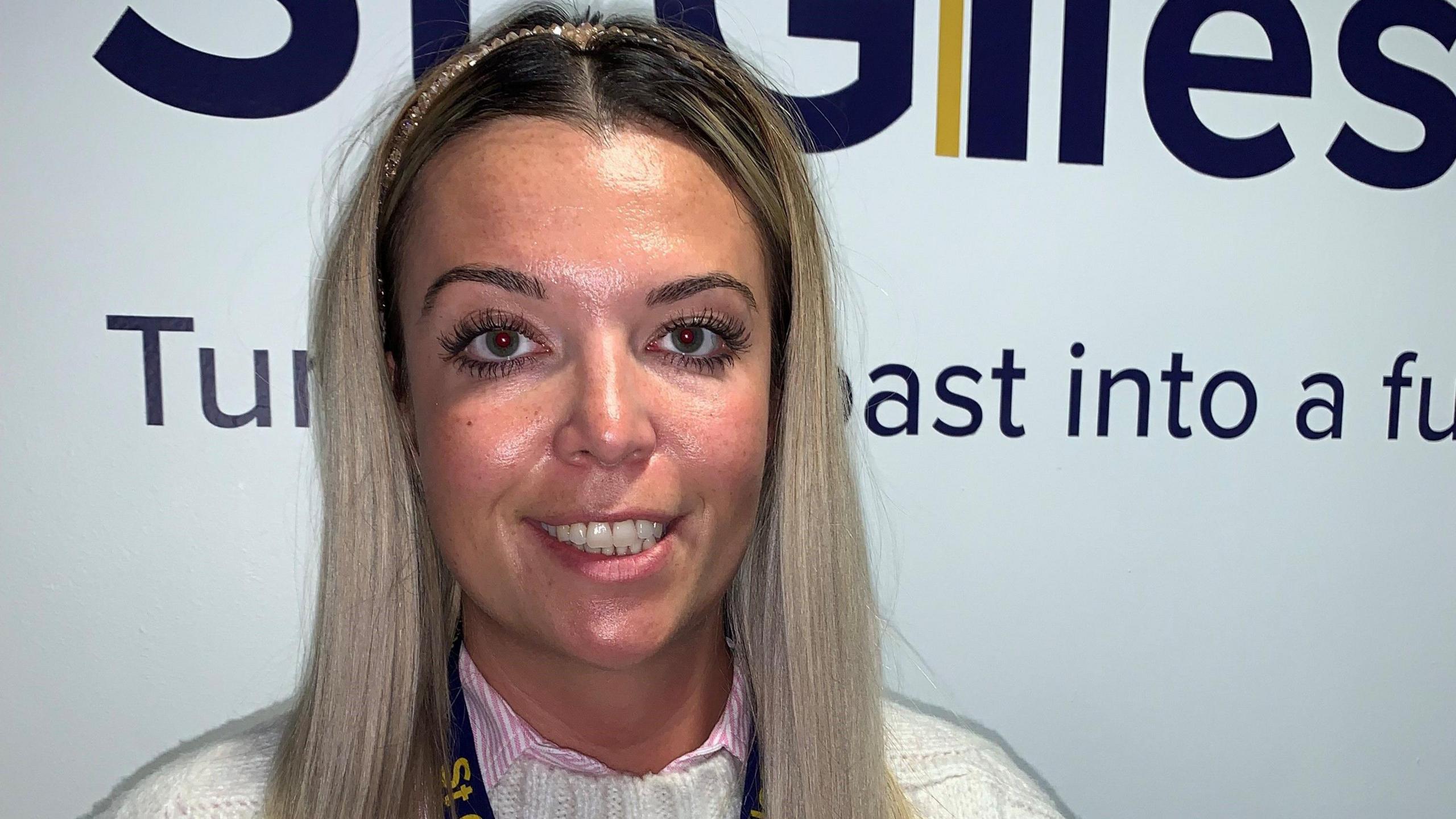 A woman with long, straight blonde hair and wearing a headband smiles. She is standing in front of a white wall that says "St Giles".