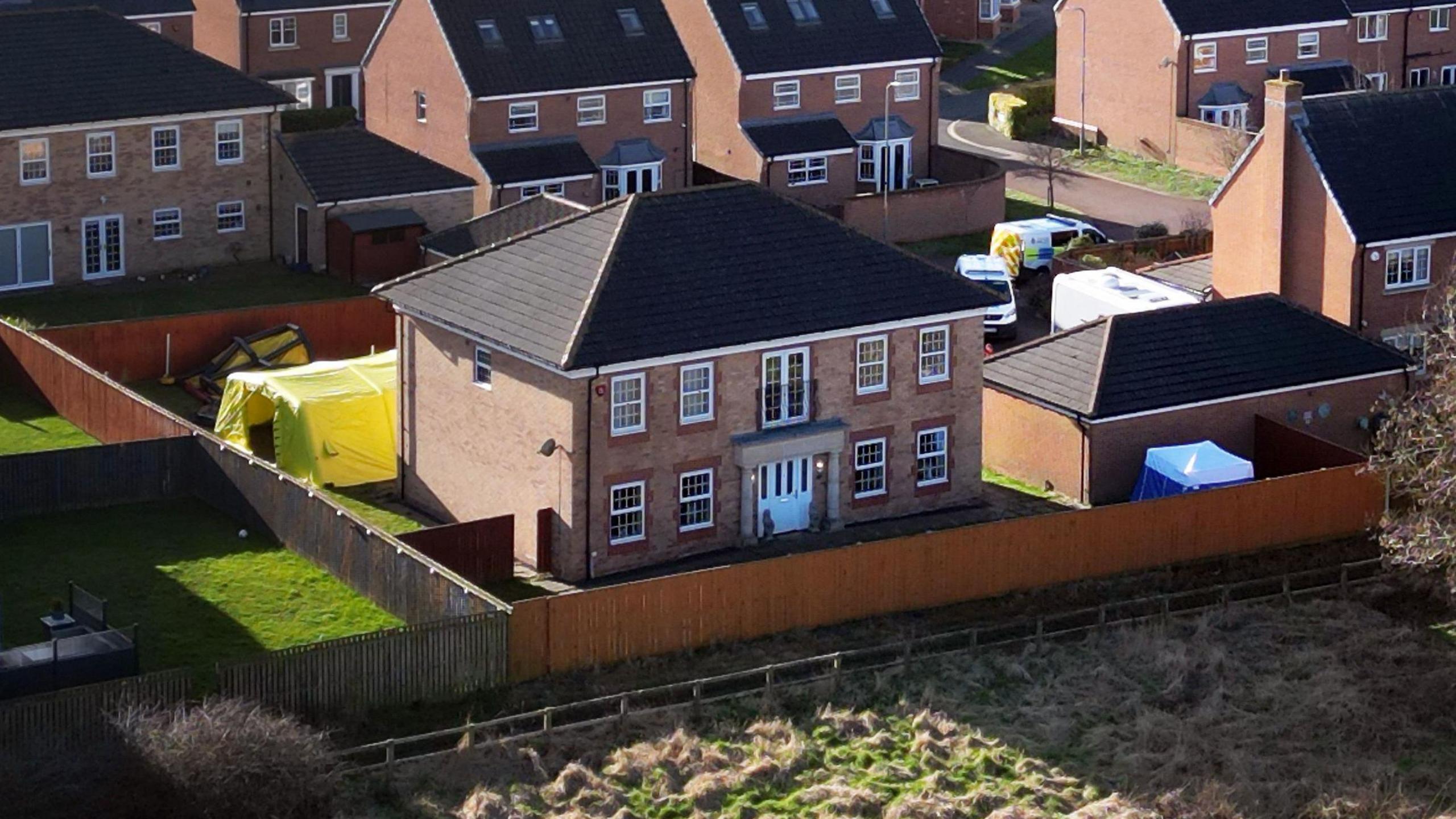 Aerial view of a large, red brick, two-storey detached house with a police forensic tent in the garden. A fence surrounds the property and other homes on the estate can be seen in the background.