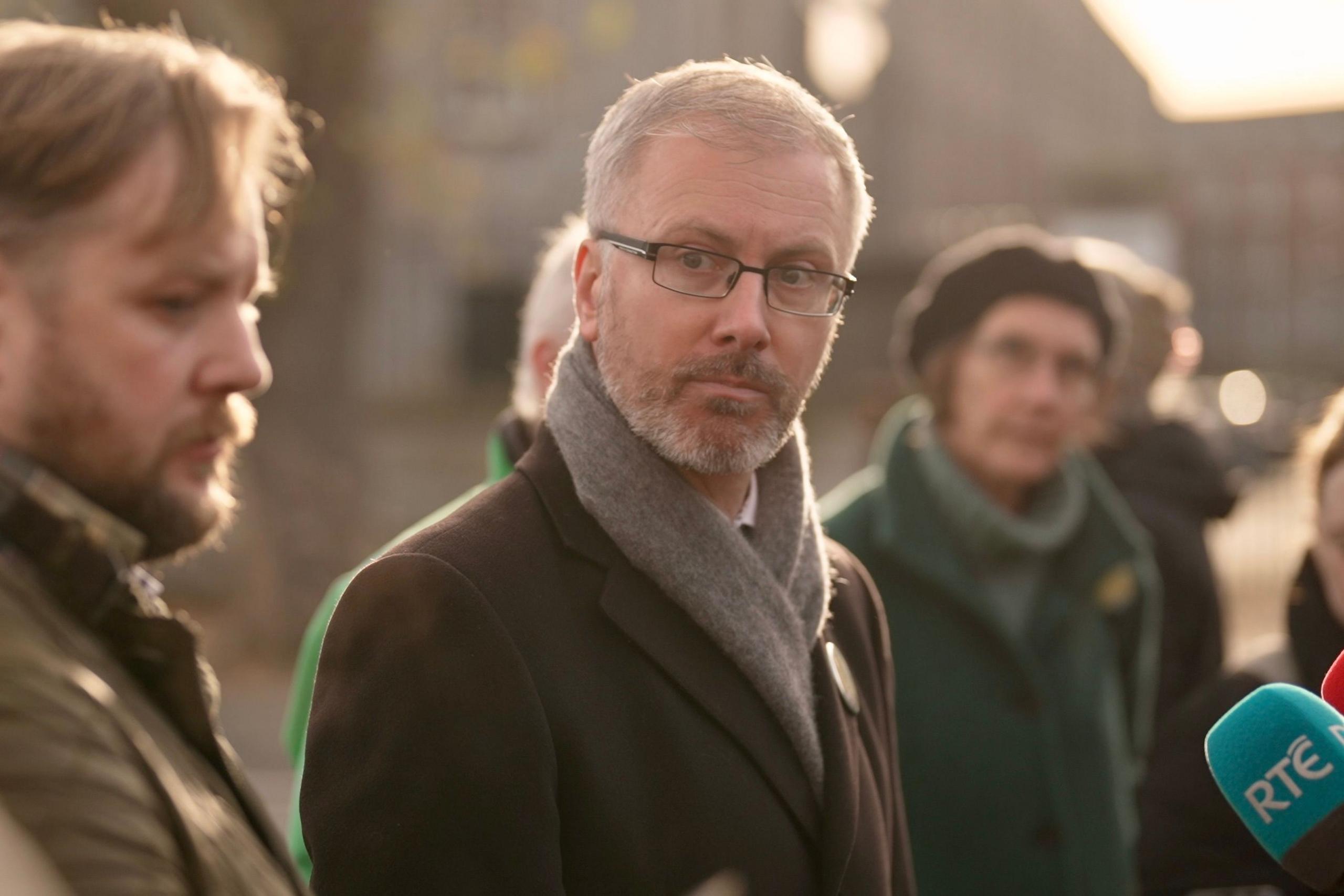 Roderic O’Gorman looks thoughtful while canvassing in the local area, an RTE - Irish radio and TV - radio microphone visible. A man and woman either side of the politician are also listening to the speaker, not pictured 