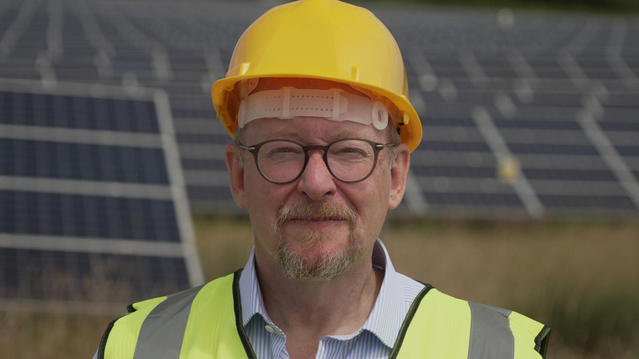 Michael Bax wearing a yellow helmet, round glasses and a high vis vest looking at the camera with a slight smile. Standing in front of rows of solar panels