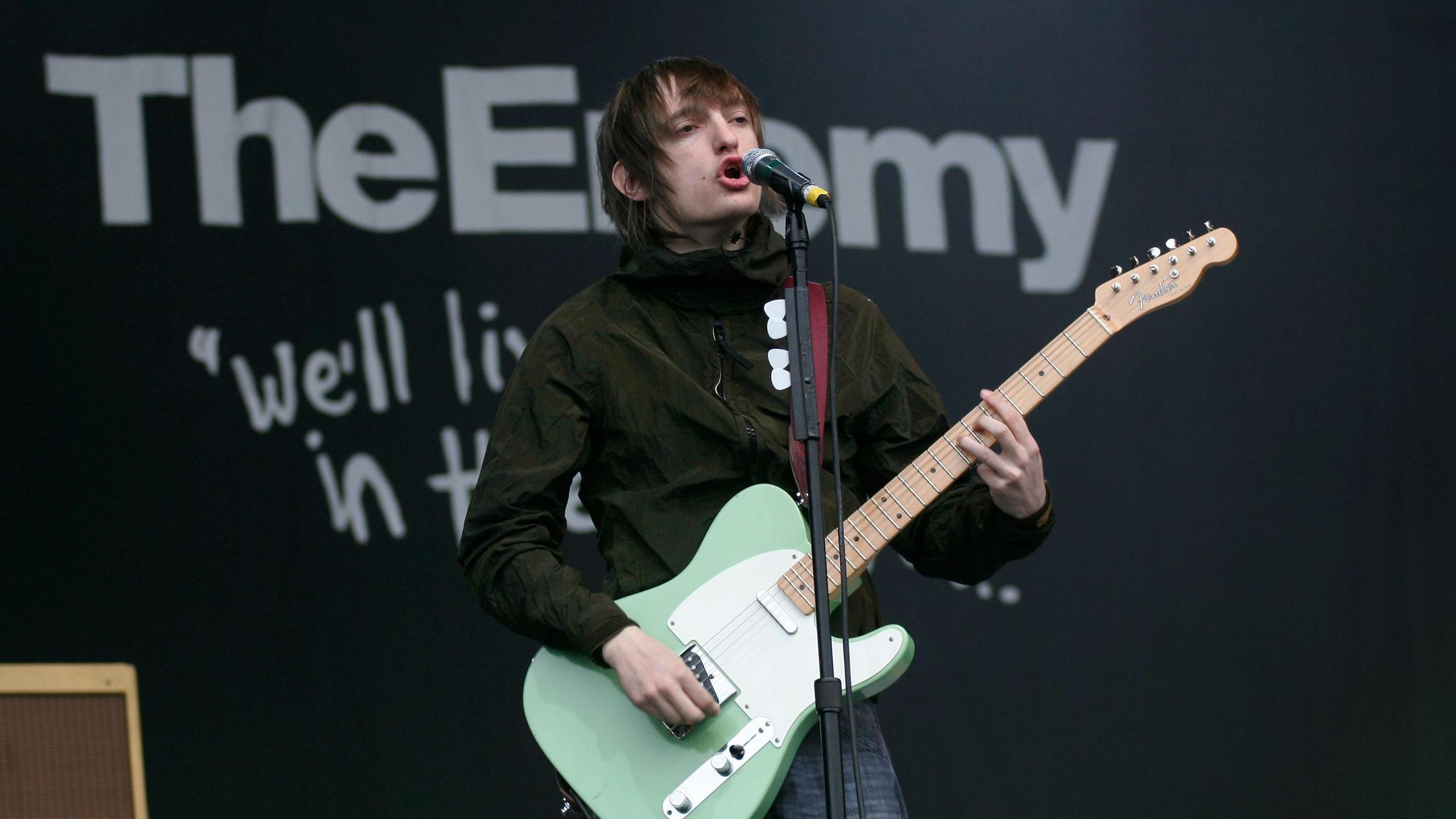 A man with mid length brown hair wearing a black jacket stands on a stage in front of a sign that says The Enemy. He is playing a green electric guitar and singing into a microphone.