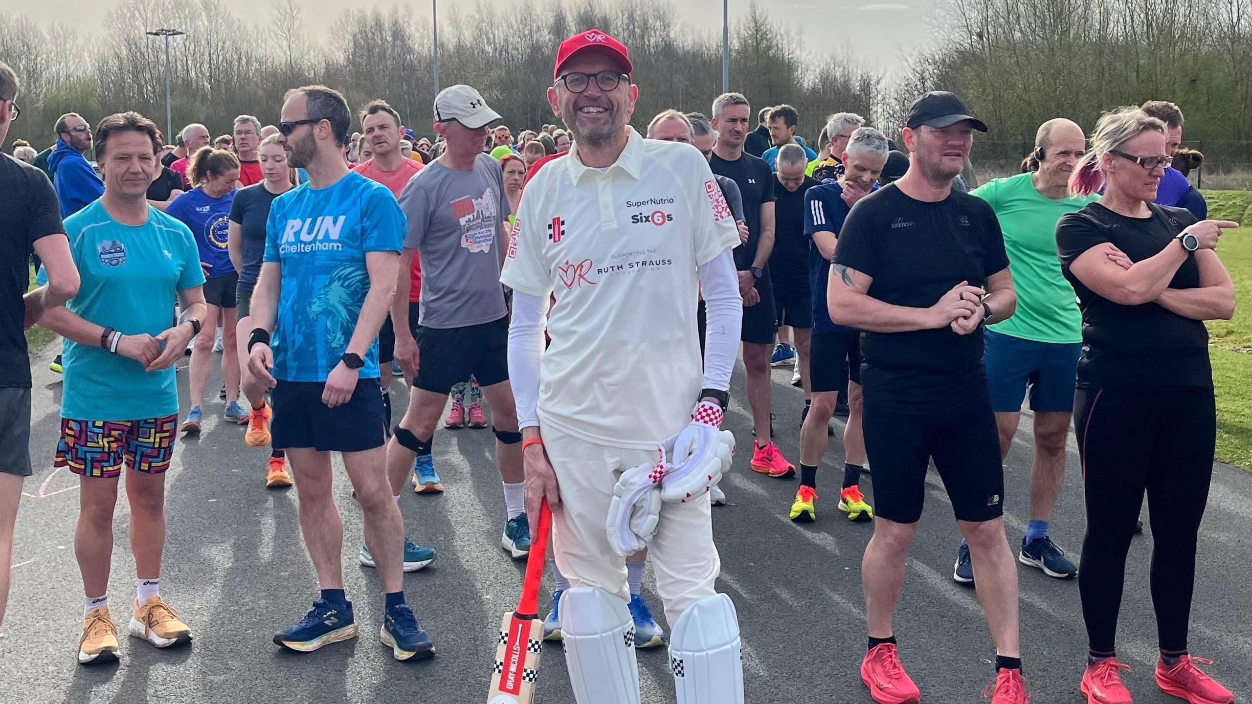A man wearing cricket whites is standing in the middle of the photo. He is wearing gloves, shin pads, and is holding a cricket bat. He has glasses and a red baseball cap on. He is surrounded by runners.