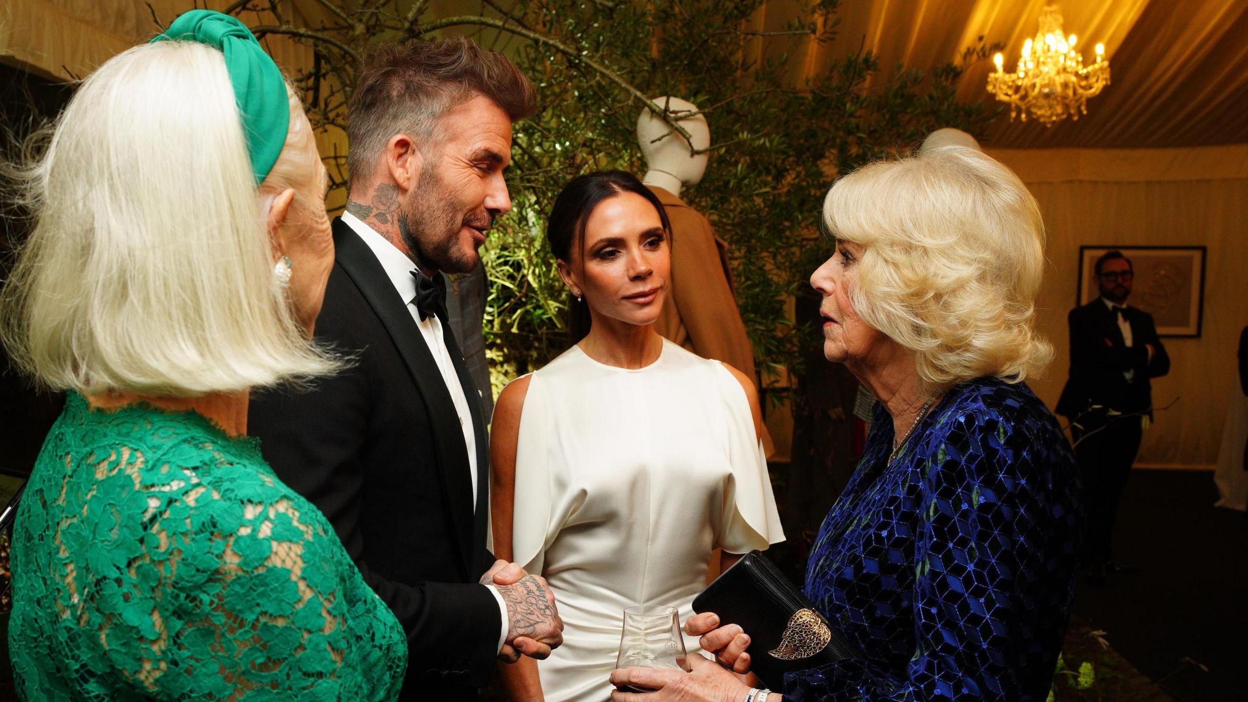 Queen Camilla stands in a huddle with the Beckhams and Dame Helen Mirren. The Queen wears a black, long-sleeved dress with a blue and green pattern. Victoria Beckham wears a sleeveless white dress with ruching at the hips. David Beckham wears a black tuxedo. Dame Helen Mirren wears a long-sleeved green dress with a lace top.