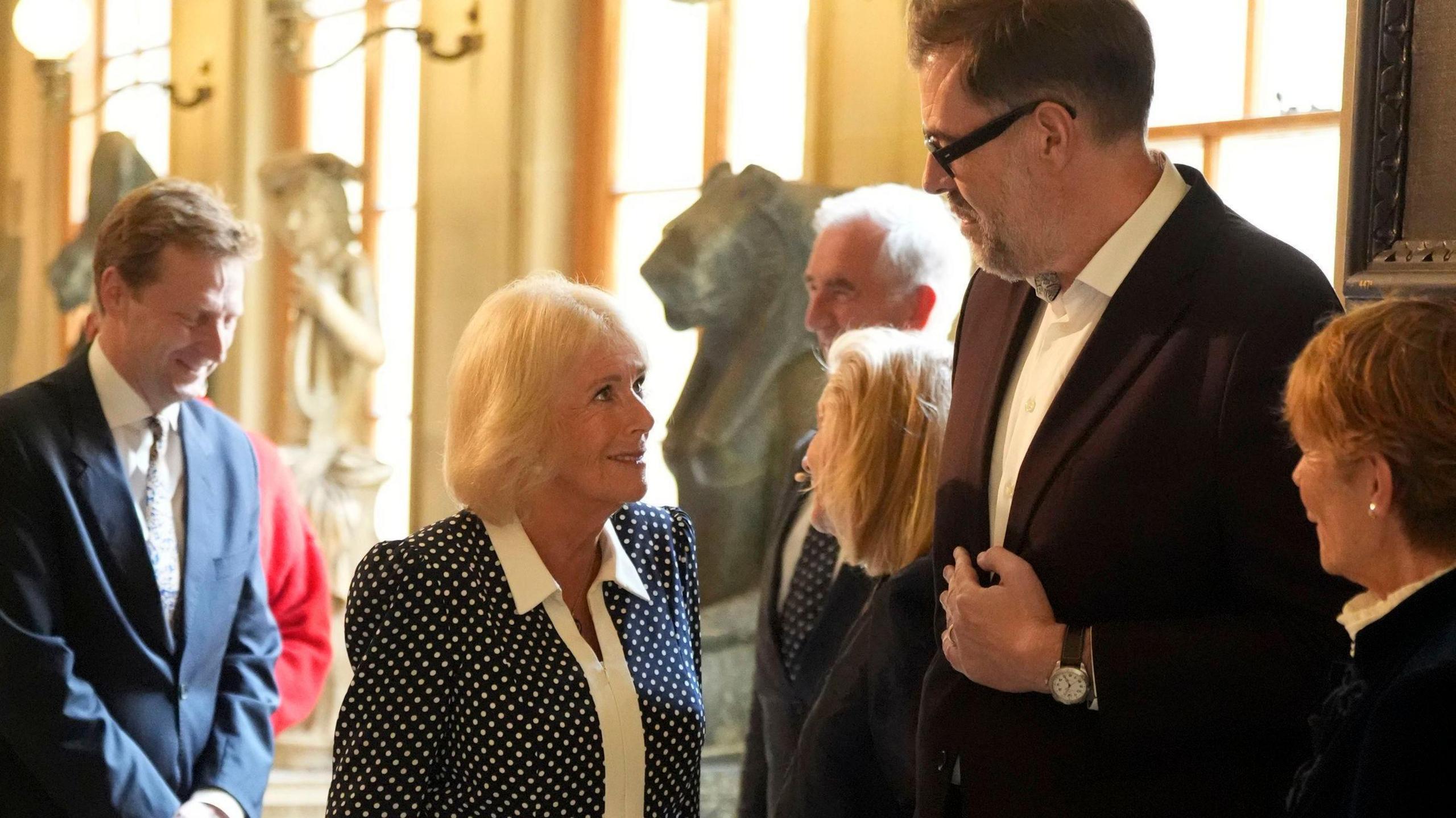 Queen Camilla meets author Richard Osman at a reception for literary figures during The Queen's Reading Room Festival at Chatsworth House. He is very tall and Queen Camilla is having to look up at him to talk.