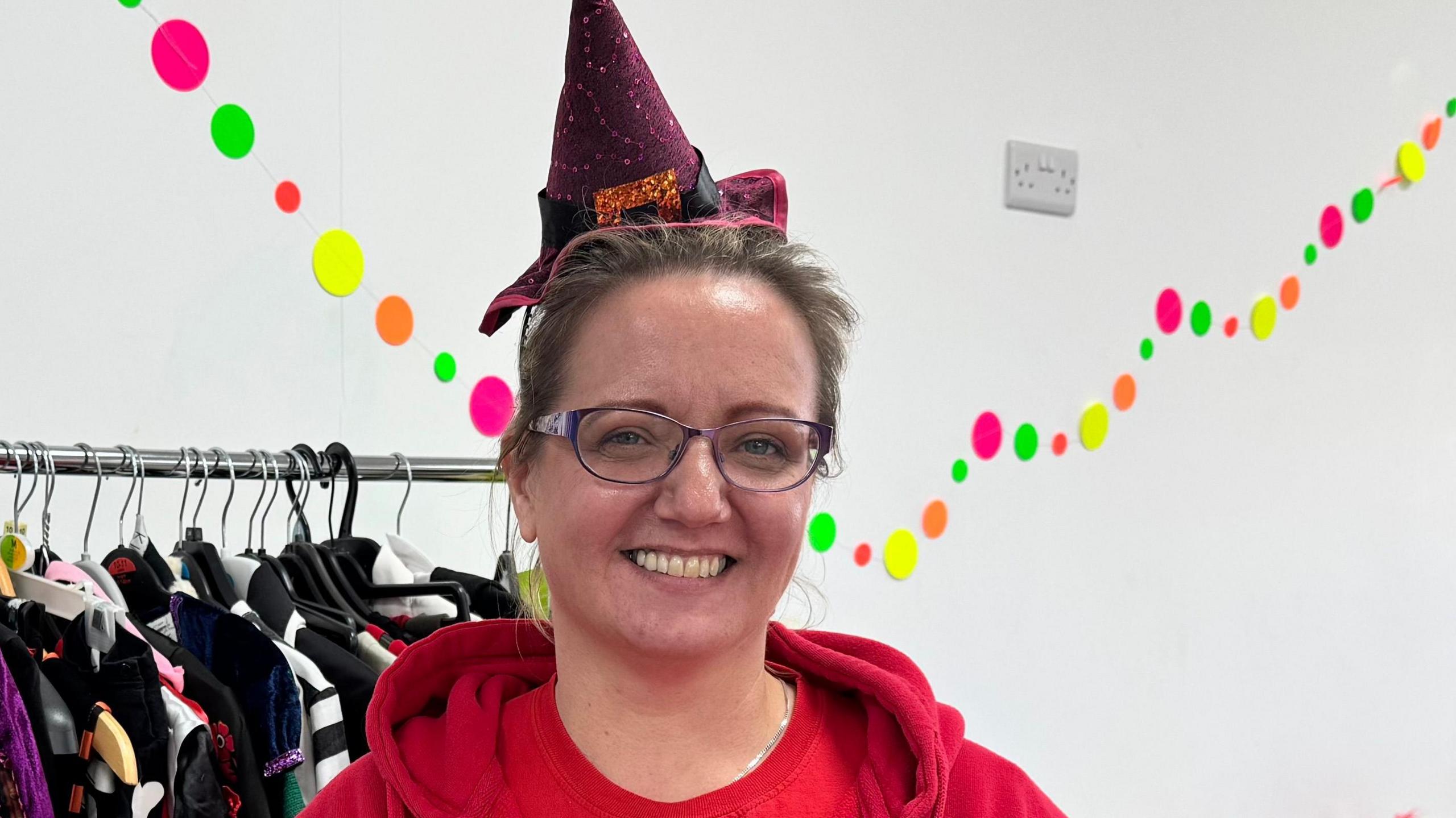 Cherry Matthews is wearing a red hoodie and a red T-shirt. She has black round glasses and brown hair. She is wearing a red spotted witch's hat with a bow on it. In the background is a rack of clothes and multi-coloured dots on the white wall. She is smiling.