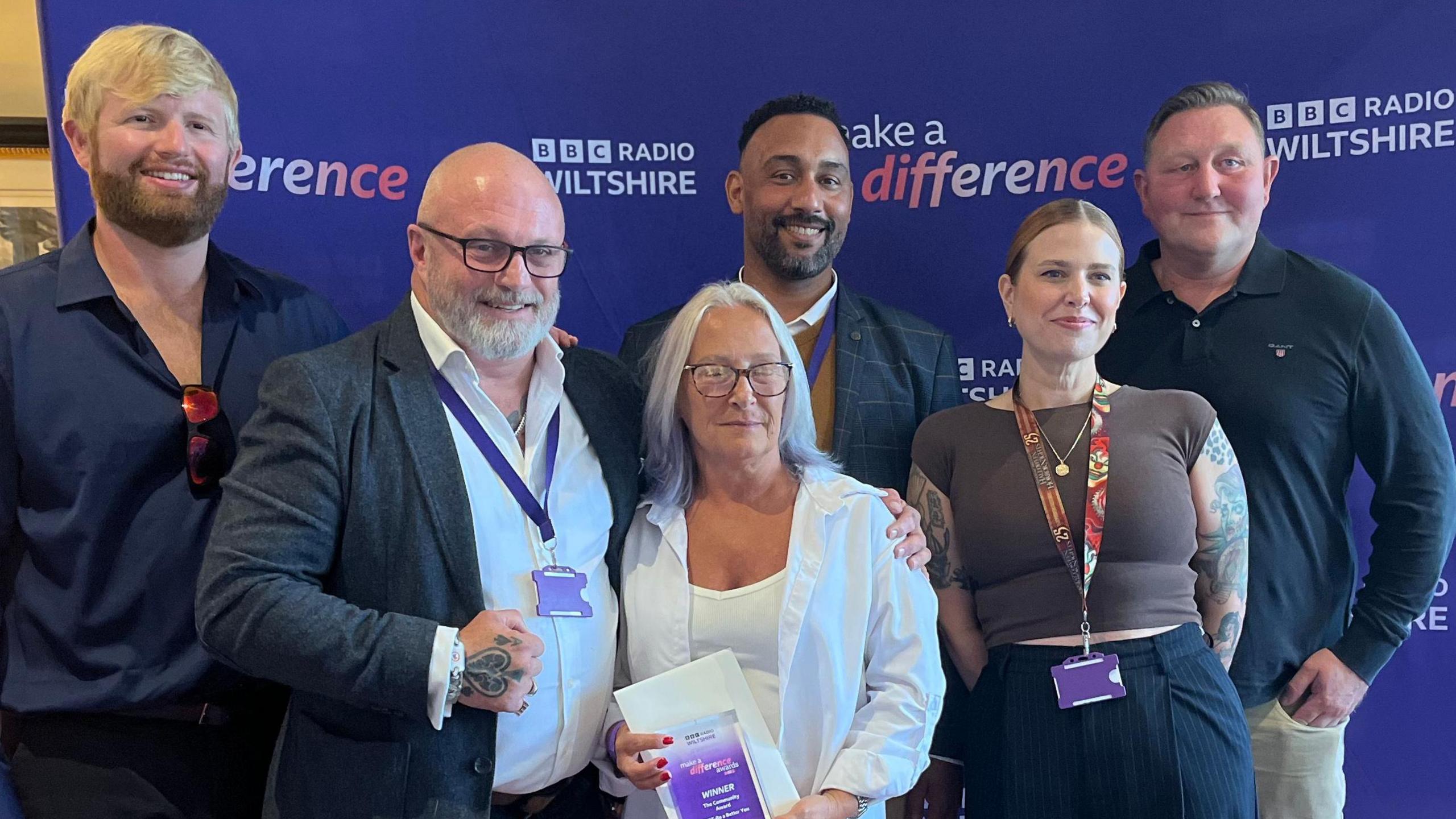 A group of people - four men, two women, stand in front of a BBC Radio Wiltshire purple banner with the woman in the middle holding the award.