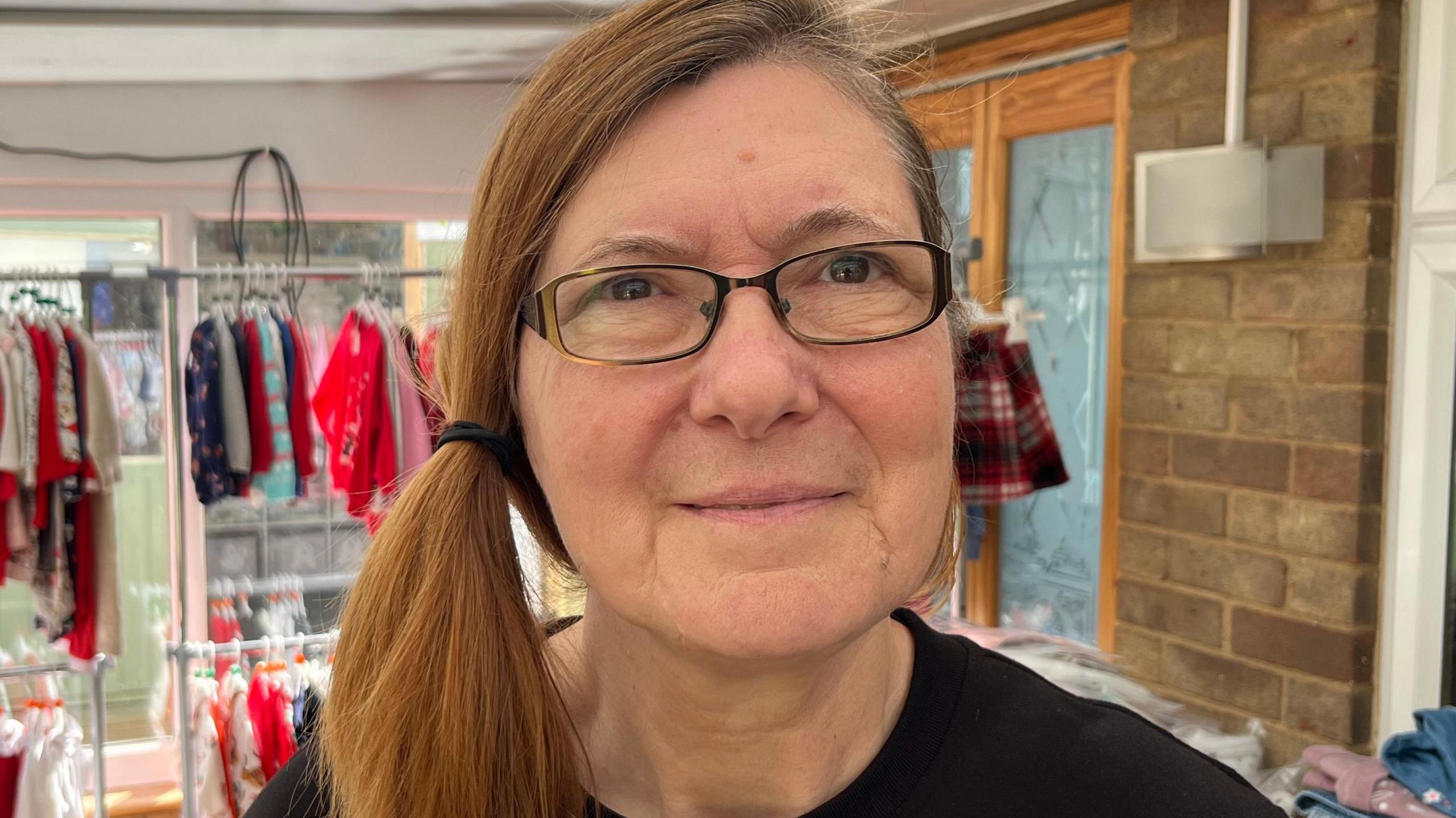Denise Martin is standing inside a room and smiling at the camera. She has ginger hair that has been put in a side ponytail to the right of her head. She is wearing rectangular framed glasses and a black top.