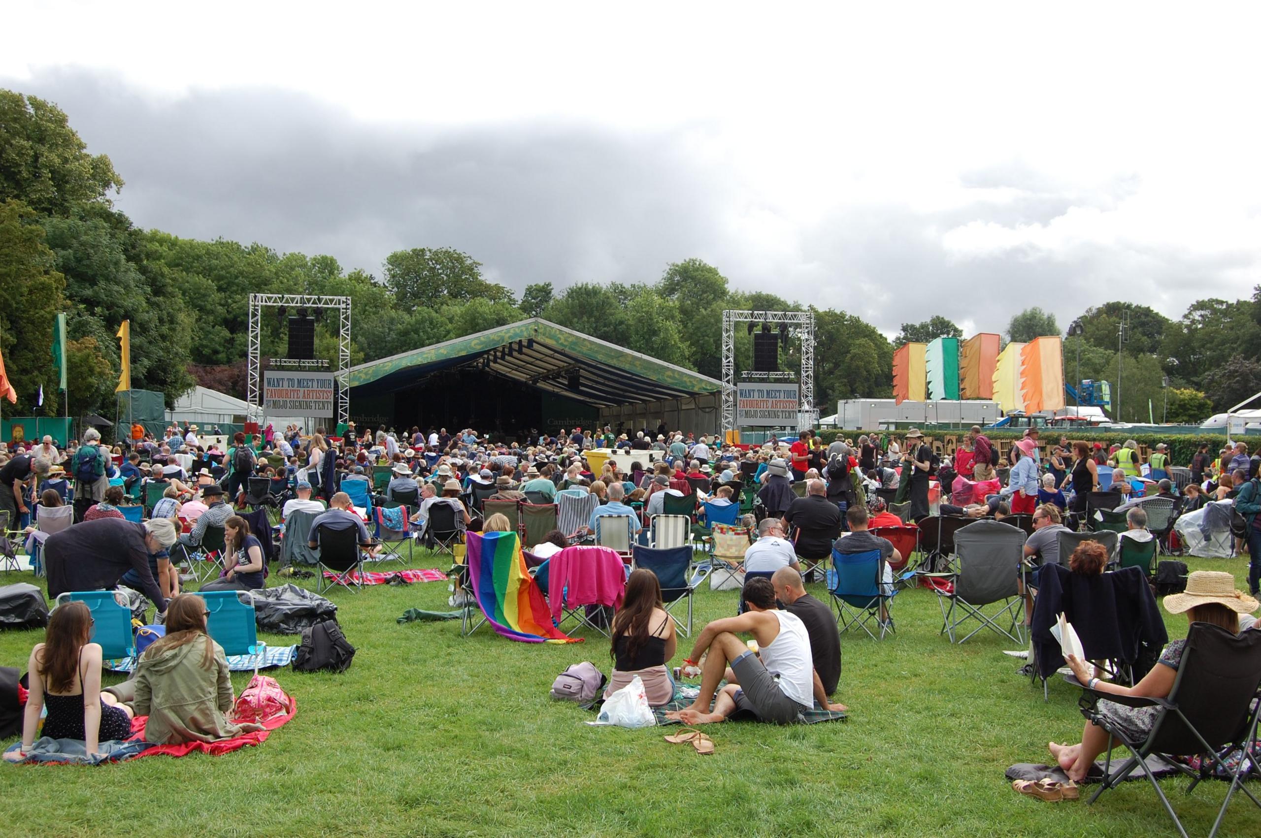 A crowd of people, many sitting on camping chairs, in front of an outdoor stage in a park. Festival flags in bright colours flutter near the stage.