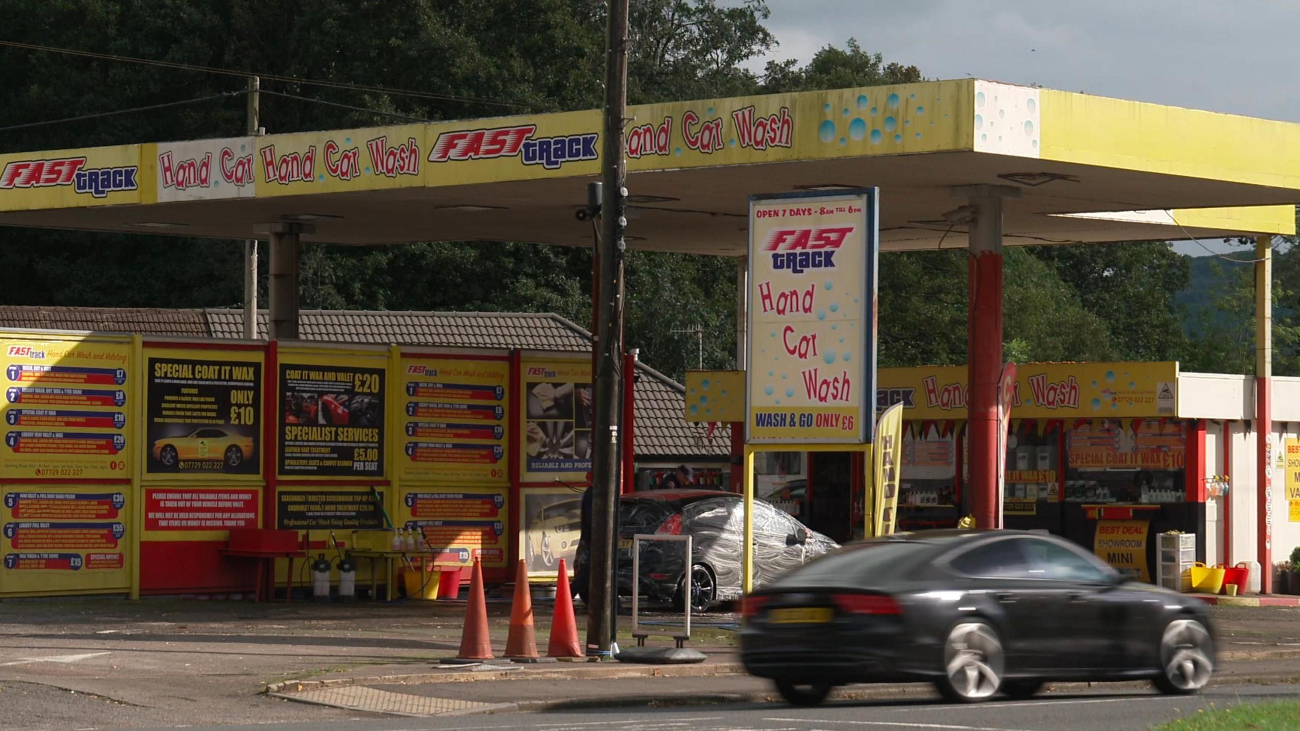 A black car is seen going past a hand car wash, which has yellow signage