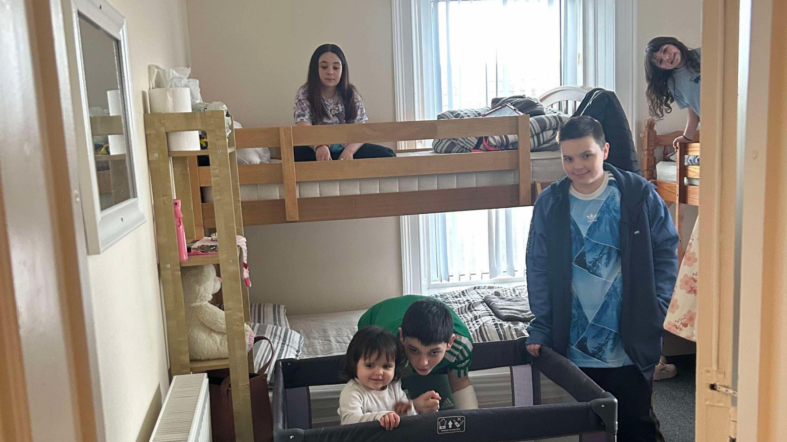 Five children of varying ages in a small bedroom. Two girls are sitting in the top bunks of bunk beds. One boy is standing next to a travel cot, another boy is bent down next to a toddler standing in the cot.