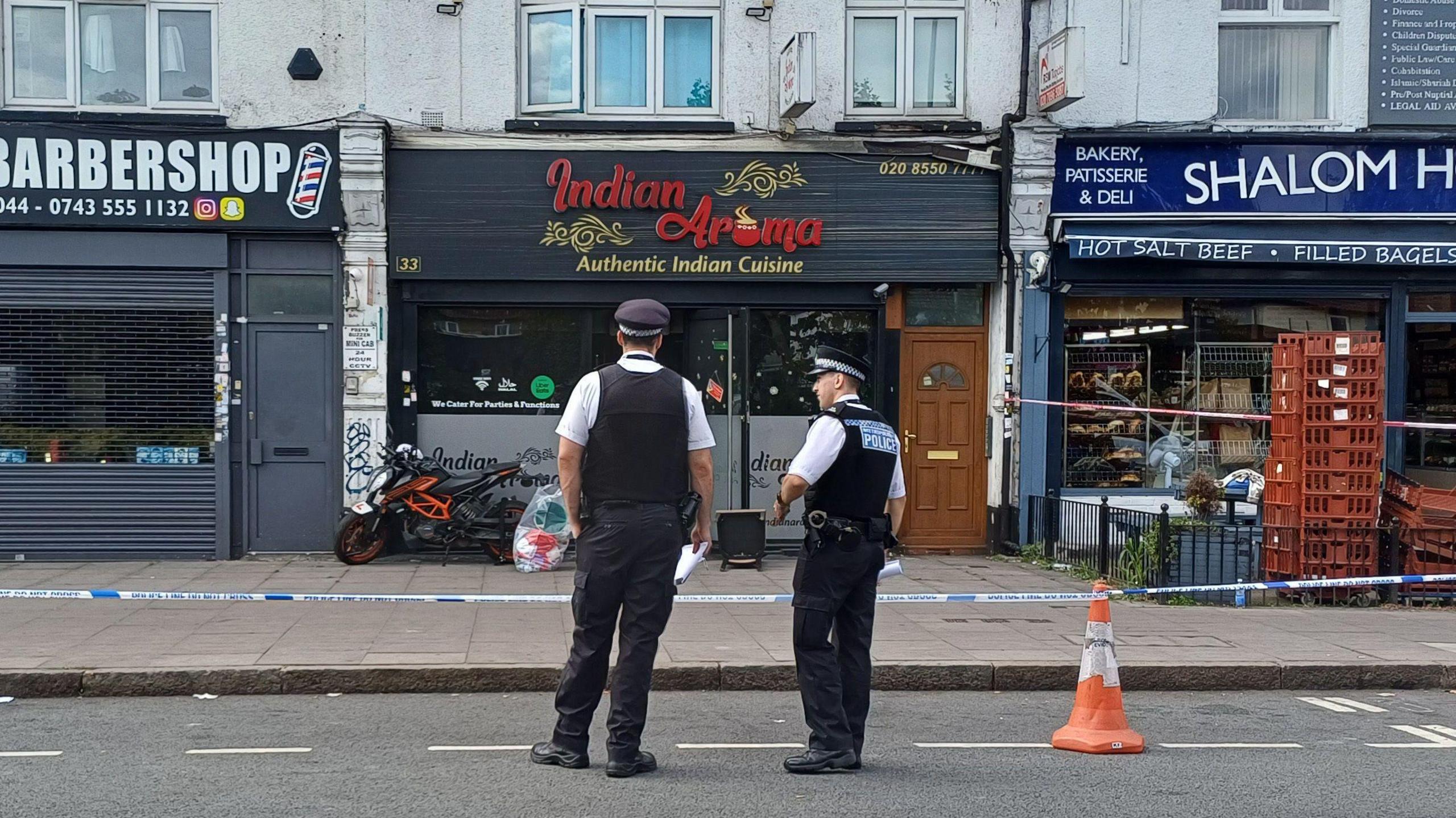 Two police officers in uniform stand on a road in front of police tape which surrounds shops and restaurants. Their backs are to the camera. Signage for the businesses pictured indicate they are a barbershop, an Indian restaurant and a deli.