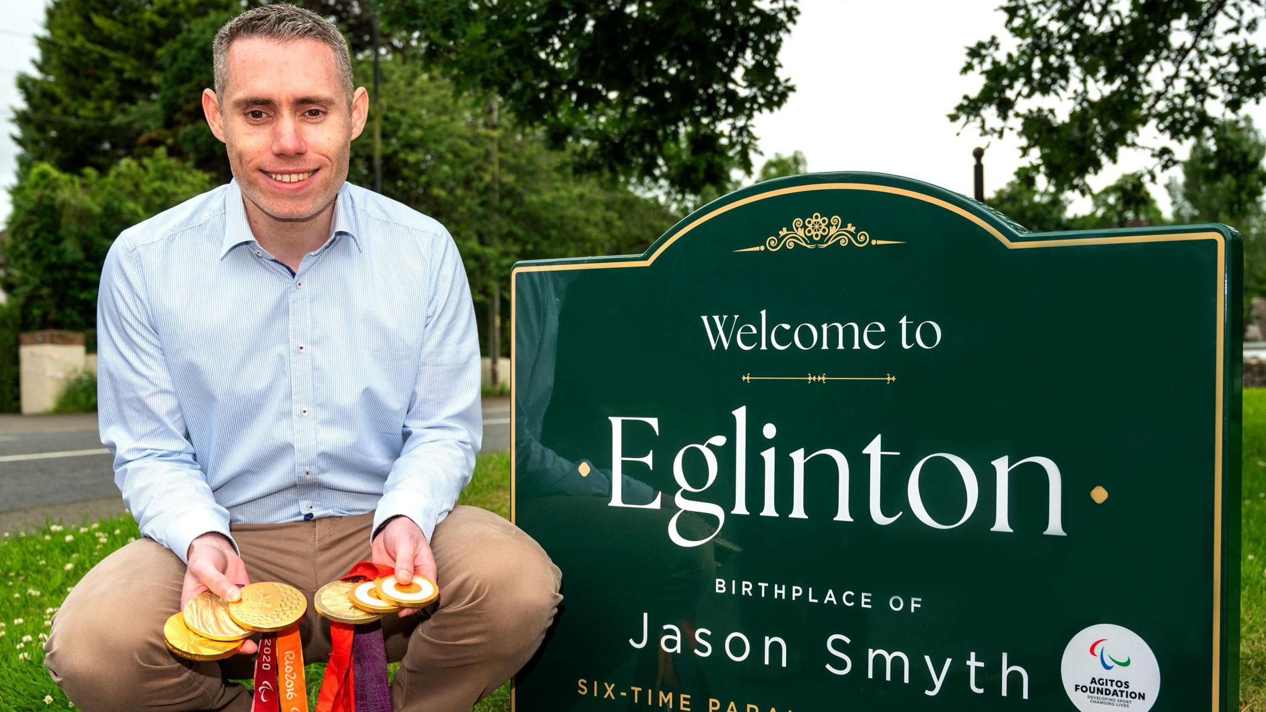 Sprinter Jason Smyth kneels beside a new welcome sign in the village where he was born. He is wearing a blue shirt and beige trousers and is holding six gold medals. He is smiling and has short greying hair.