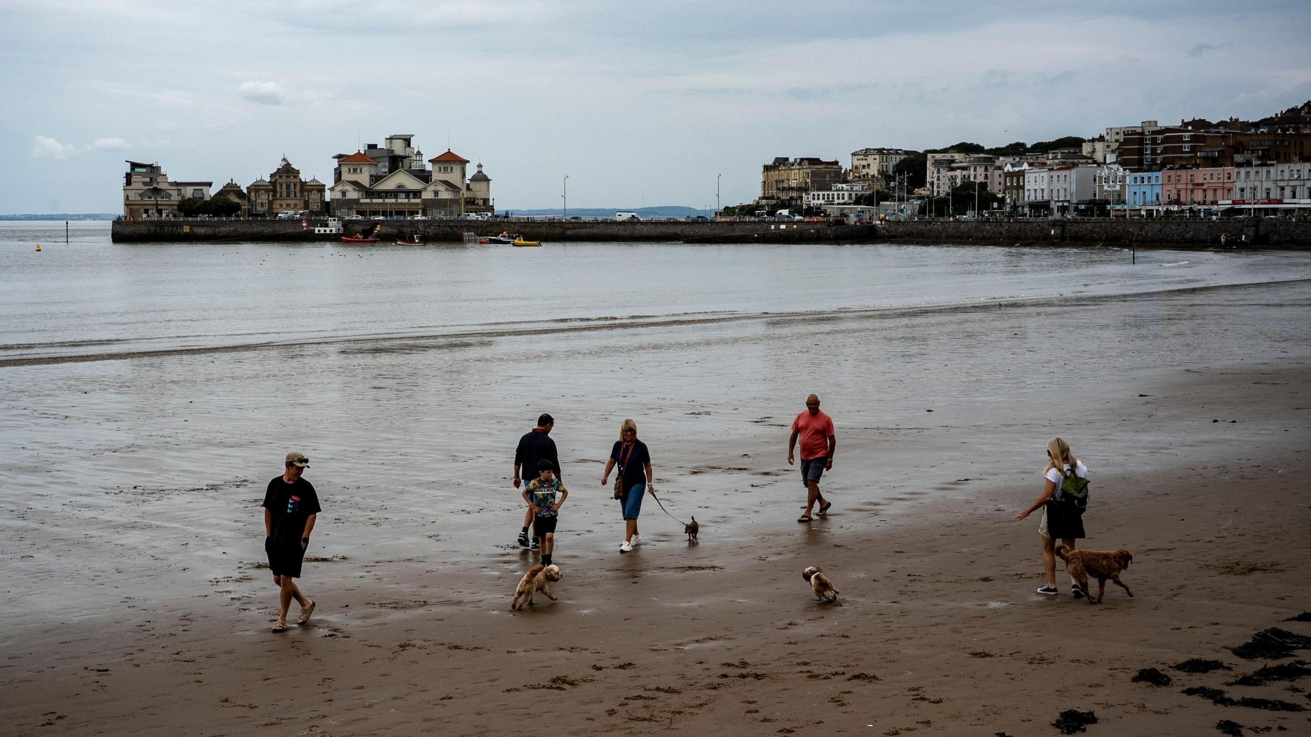 Overcast skies on Weston-super-Mare beach where five dog walkers and beach-goers walk along the sand. Housing and other buildings are visible in the background