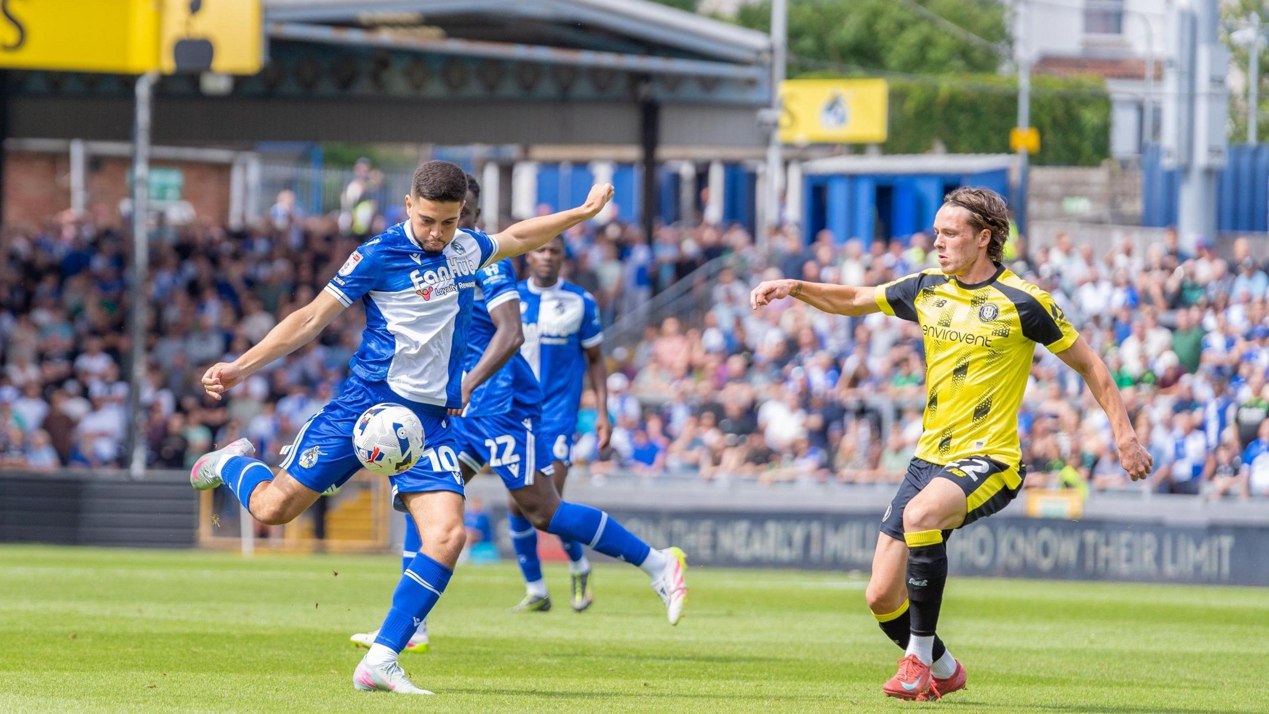 A player from Bristol Rovers, in the club's traditional blue and white quarters kit, shoots during the match with Harrogate Town at the Memorial Stadium. A Harrogate player in a yellow top and dark shorts is trying to block the shot