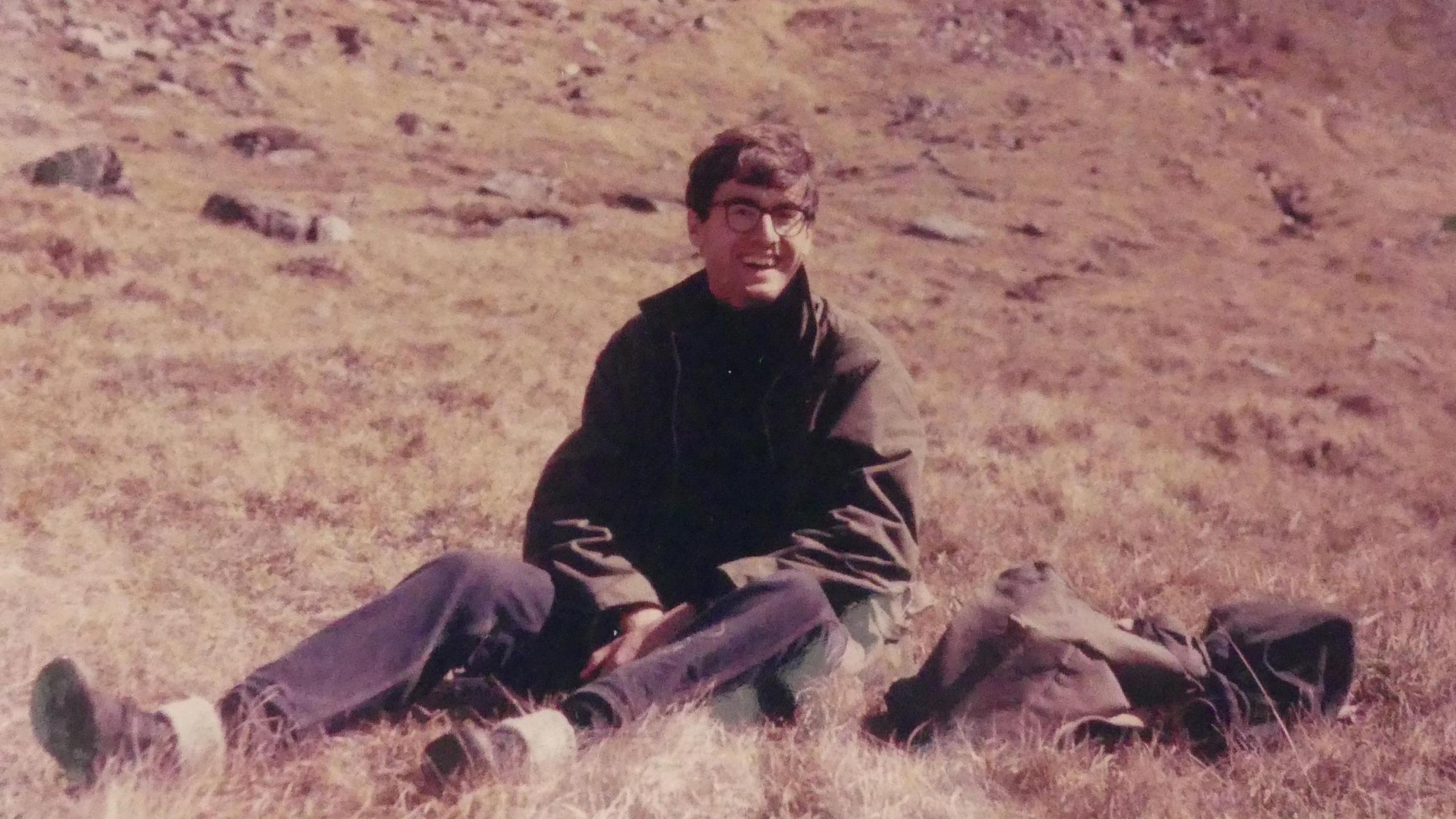 Nigel is smiling as he sits on moorland. There are rocks in the distance. He is wearing walking boots and long trousers and a long-sleeved jacket. There is a bag beside him.