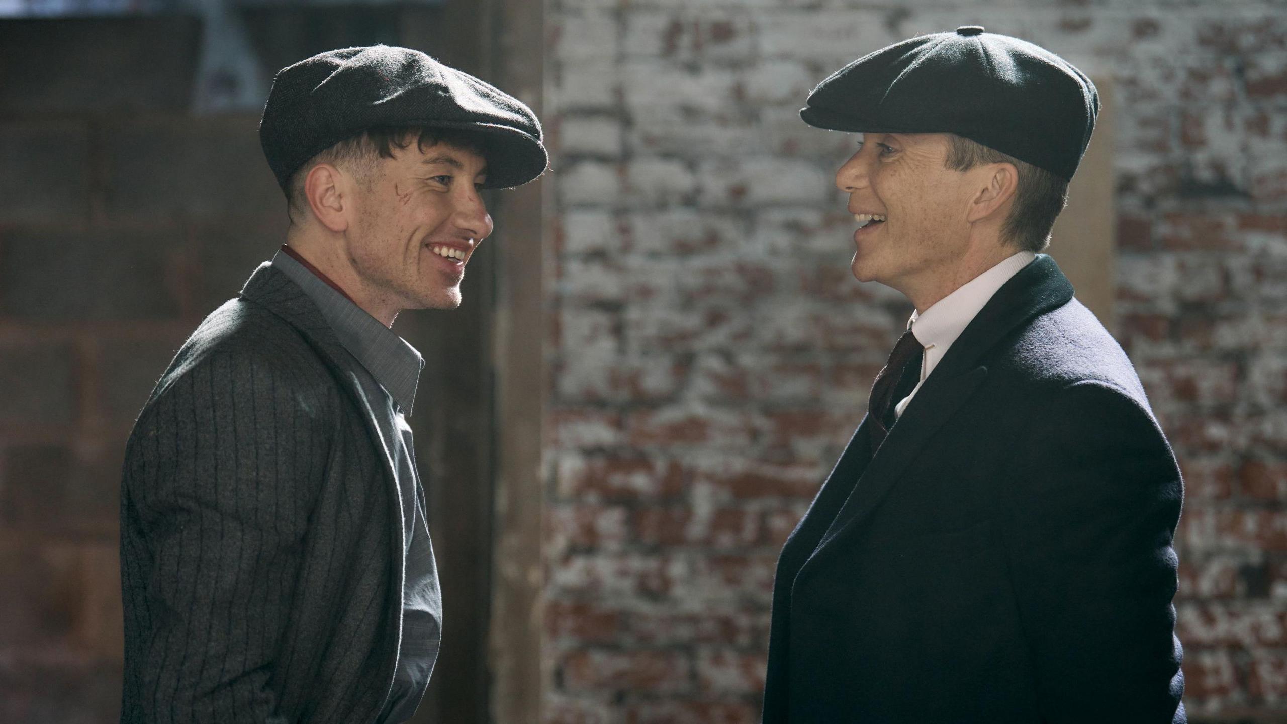 Barry Keoghan and Cillian Murphy, in the upcoming Peaky Blinders film. They are laughing and both are wearing dark suits and caps.