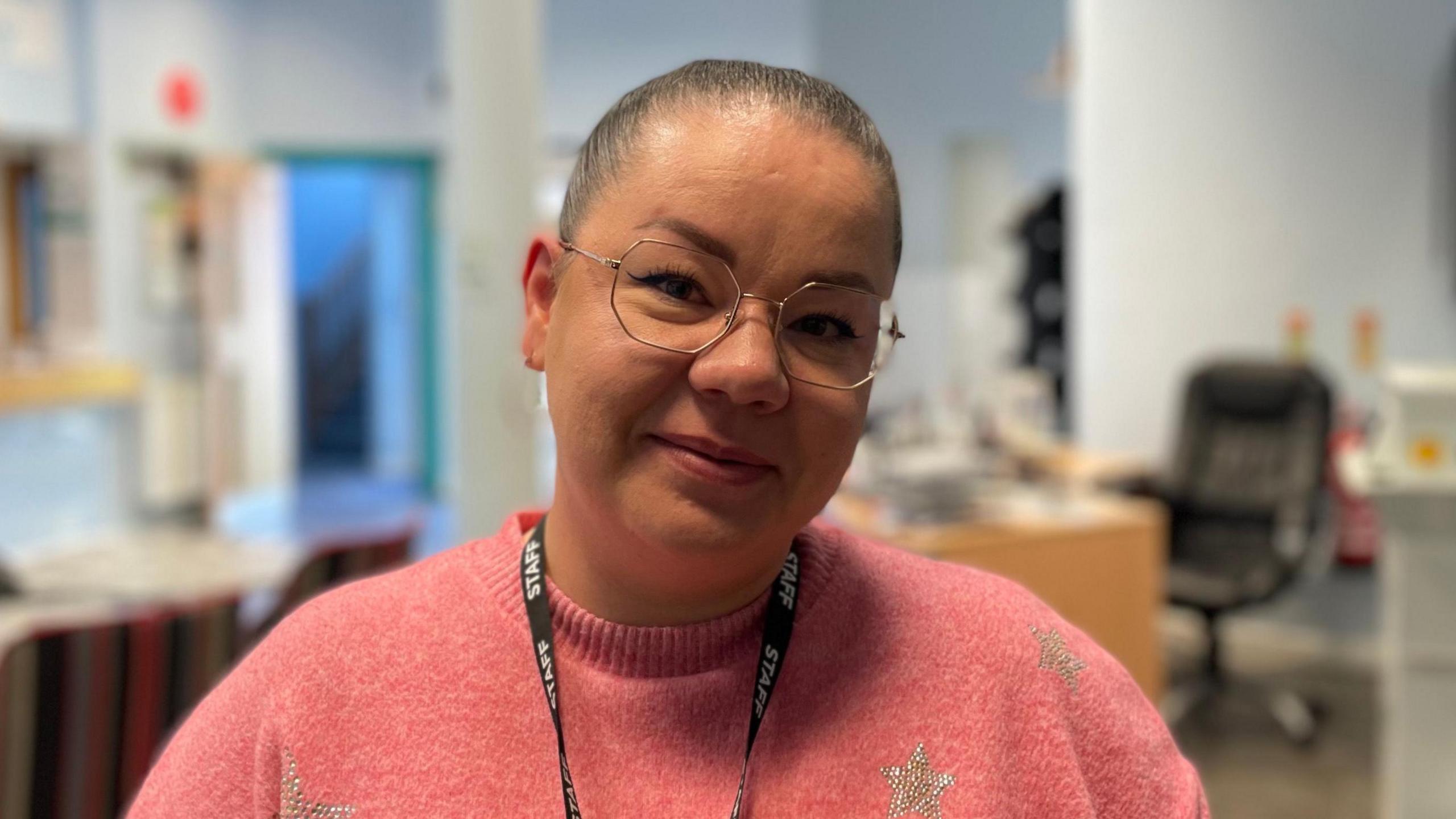 Simona Cismasu in the lobby of the Rosmini centre. There is a reception area in the background. She has tied back dark hair and glasses. She is wearing a pink jumper with stars on it and she has a black lanyard around her neck.
