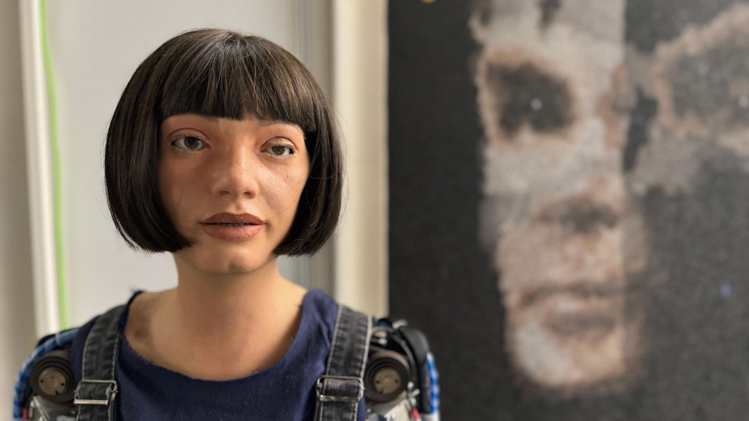 Humanoid robot with a black bob haircut and wearing denim dungarees.