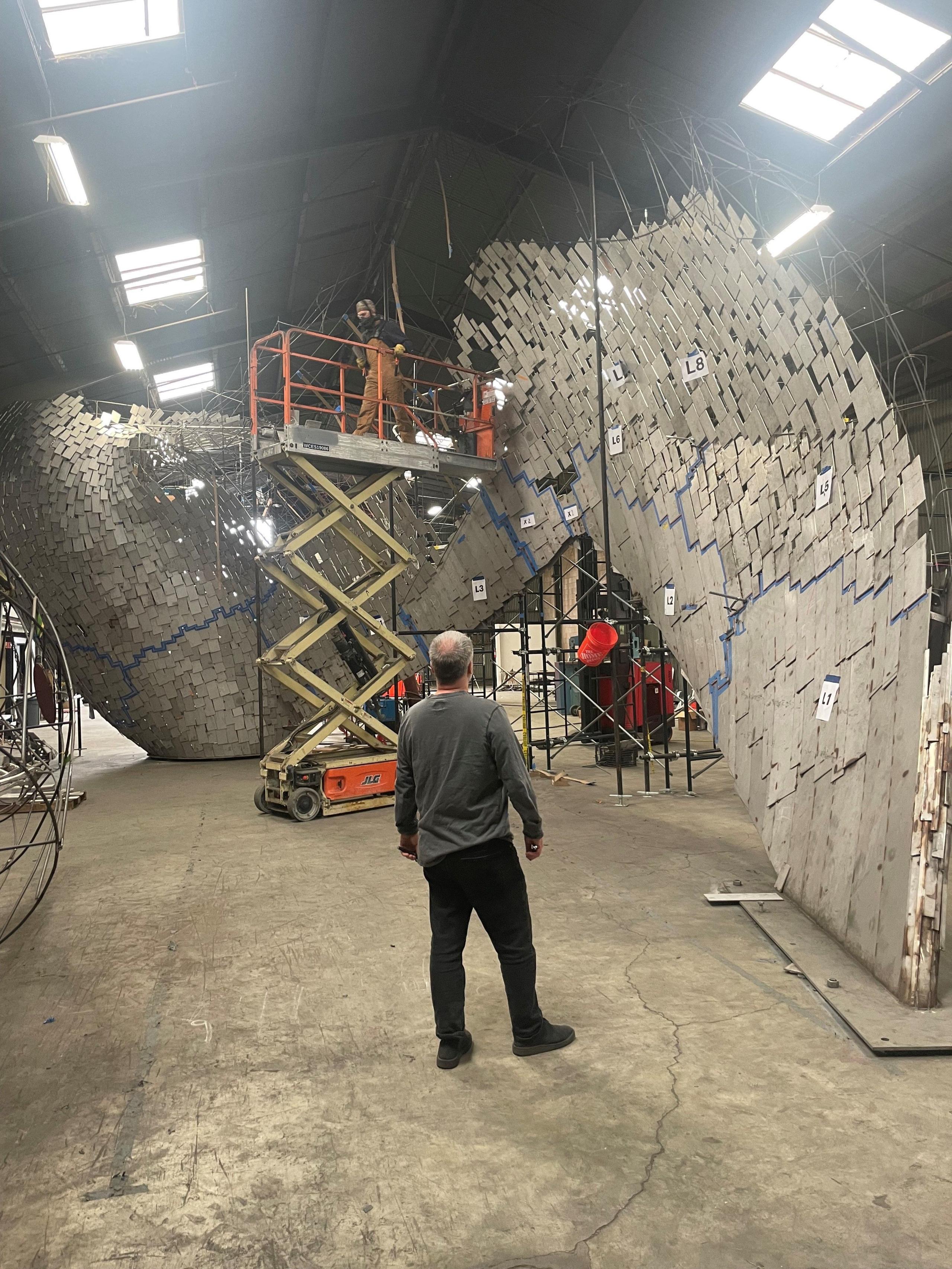 Andy Scott photographed from behind, viewing the construction process of the loon bird sculpture, 'The Calling', in a high ceilinged warehouse 