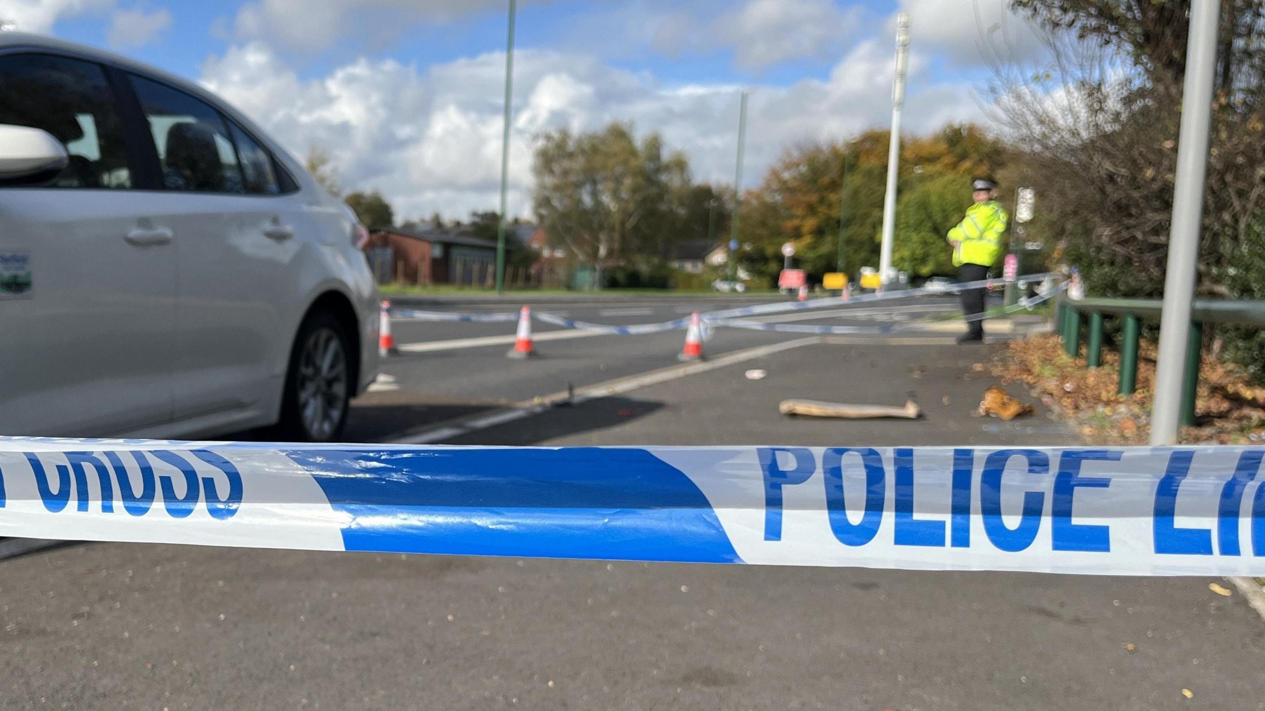 A white taxi inside a police cordon which is visible in the foreground. A police officer is stood in the background.