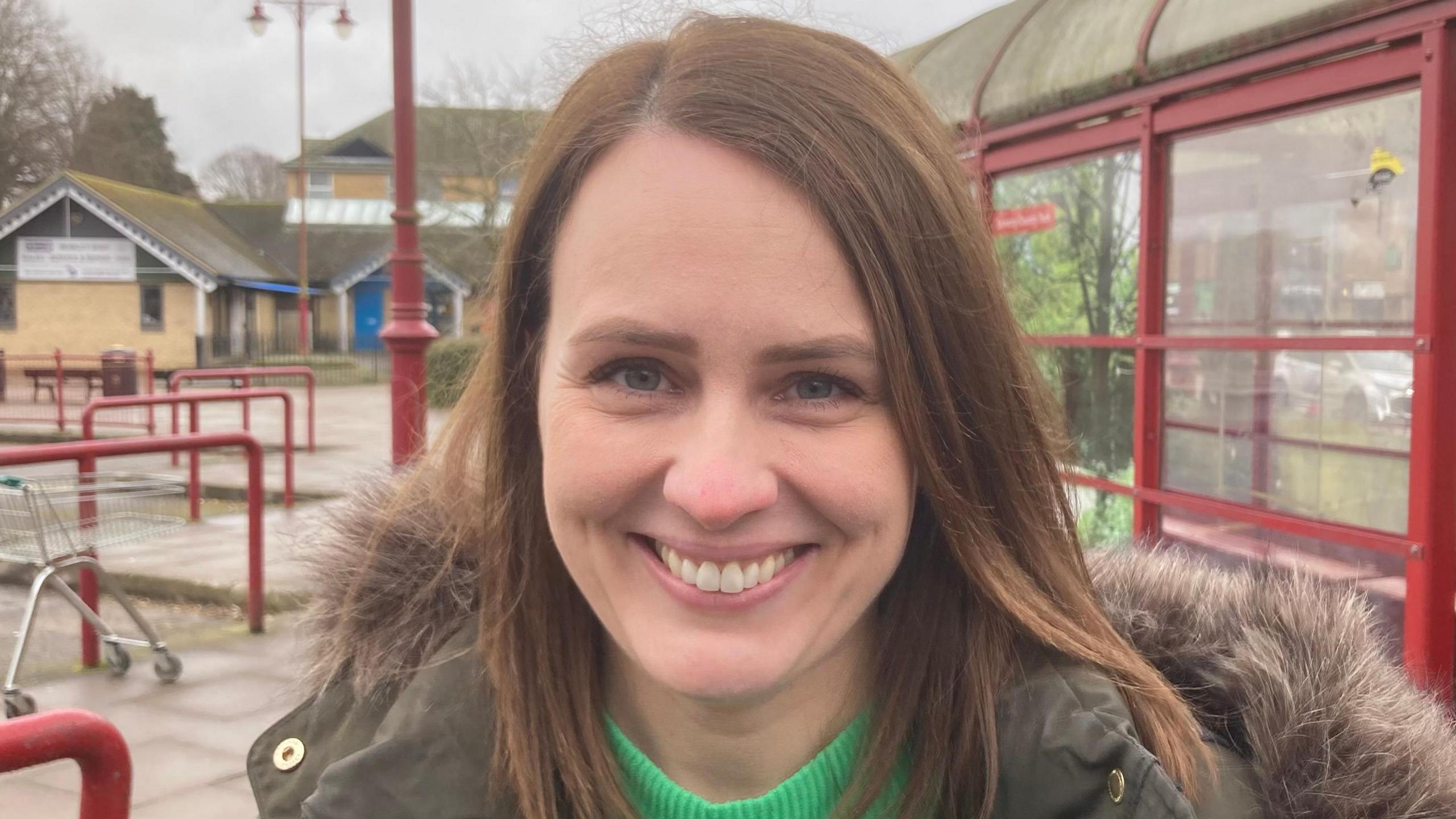 Hayley Charlesworth with long brown hair smiling at the camera. She is wearing a green coat with a furry hood and a light green top. There is a red-framed glass bus shelter behind her. There is a shopping trolley to the left.