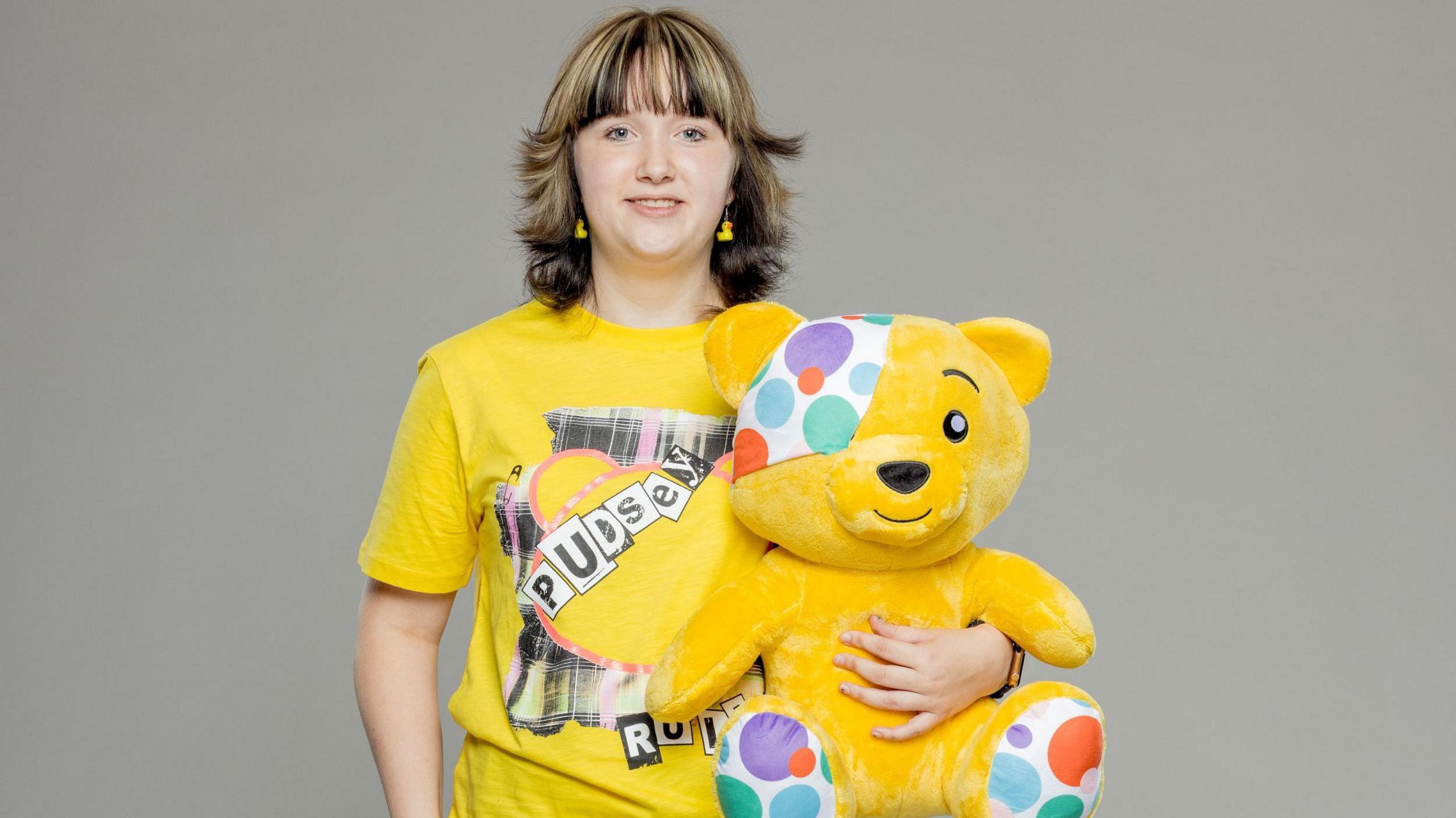 Eloise, a teenager with highlighted brown and blonde hair, hugs a Pudsey Bear soft toy while wearing a matching yellow T-shirt with the bear's bead on it.