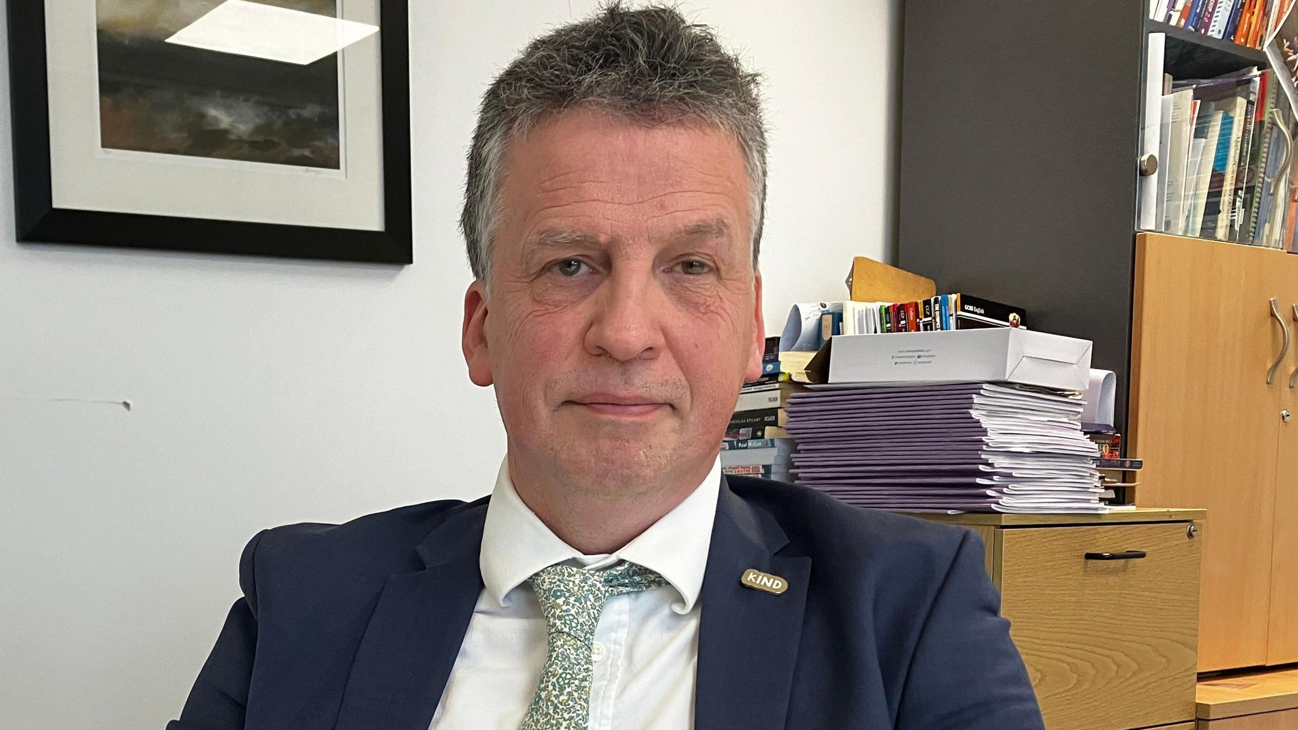 Nigel Youngman sits in a chair in front of some cabinets. He has grey hair and is wearing a navy suit, white shirt and green floral tie.