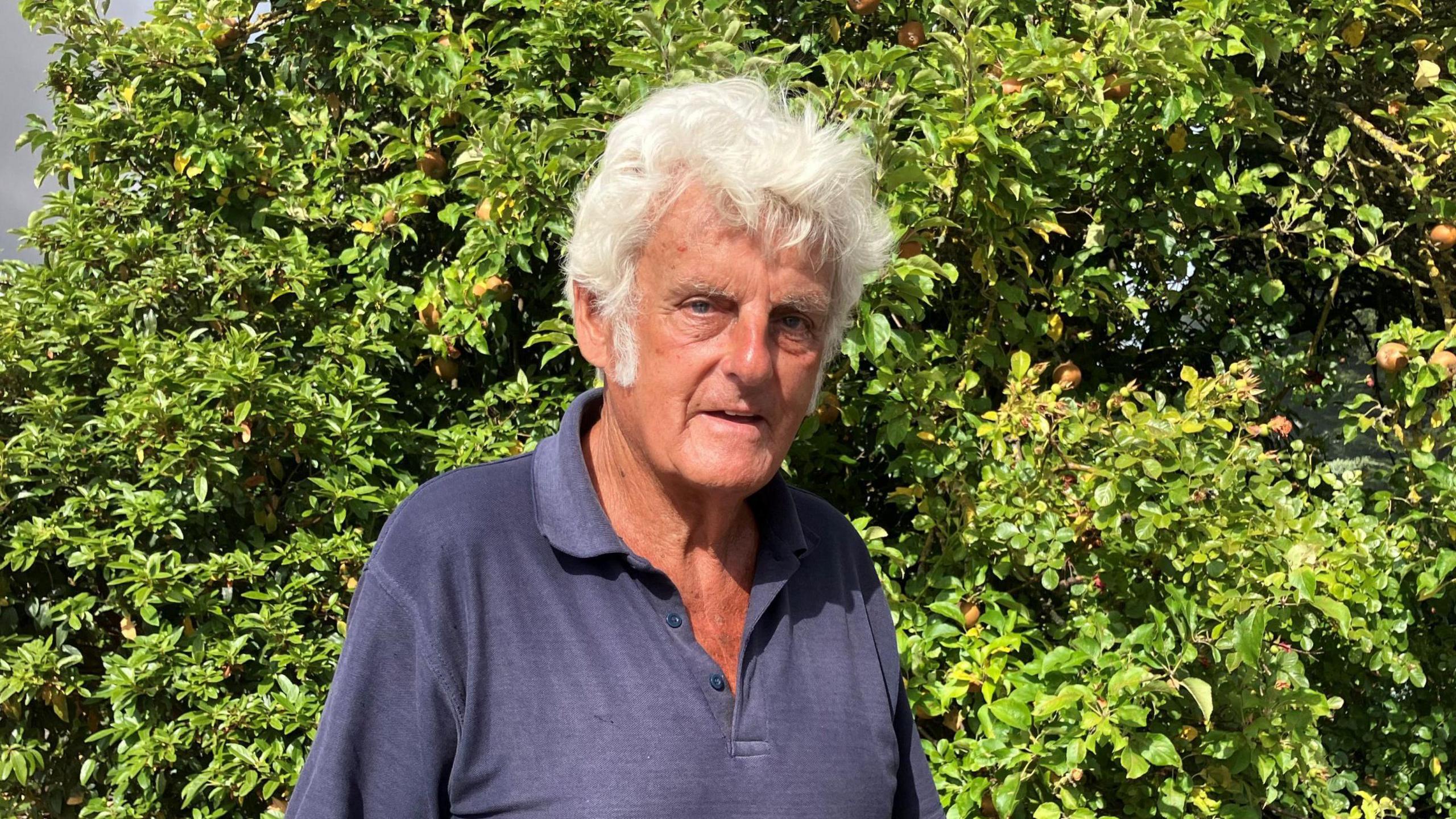 A elderly man wearing a dark blue polo shirt and looking at the camera. He is standing in front of a green bush