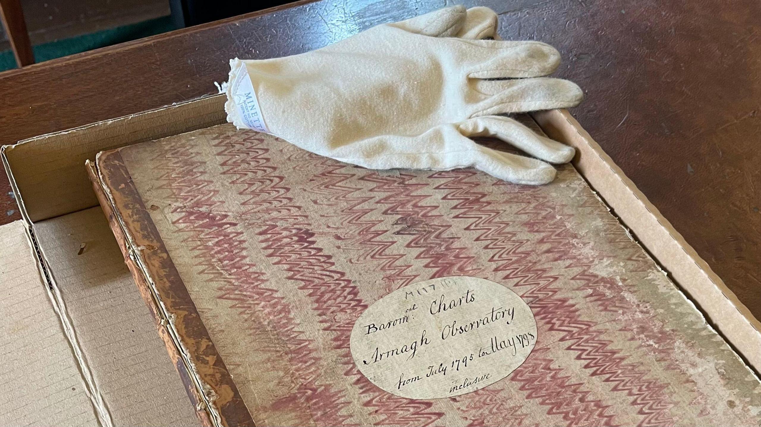 A close-up of a very old book containing records of weather observations from Armagh Observatory from July 1795 to May 1798. The book, which has a hand-written cover and is decorated with wavy red lines, is in a cardboard box on a wooden desk. A white glove used for handling the delicate paper is lying on the book.
