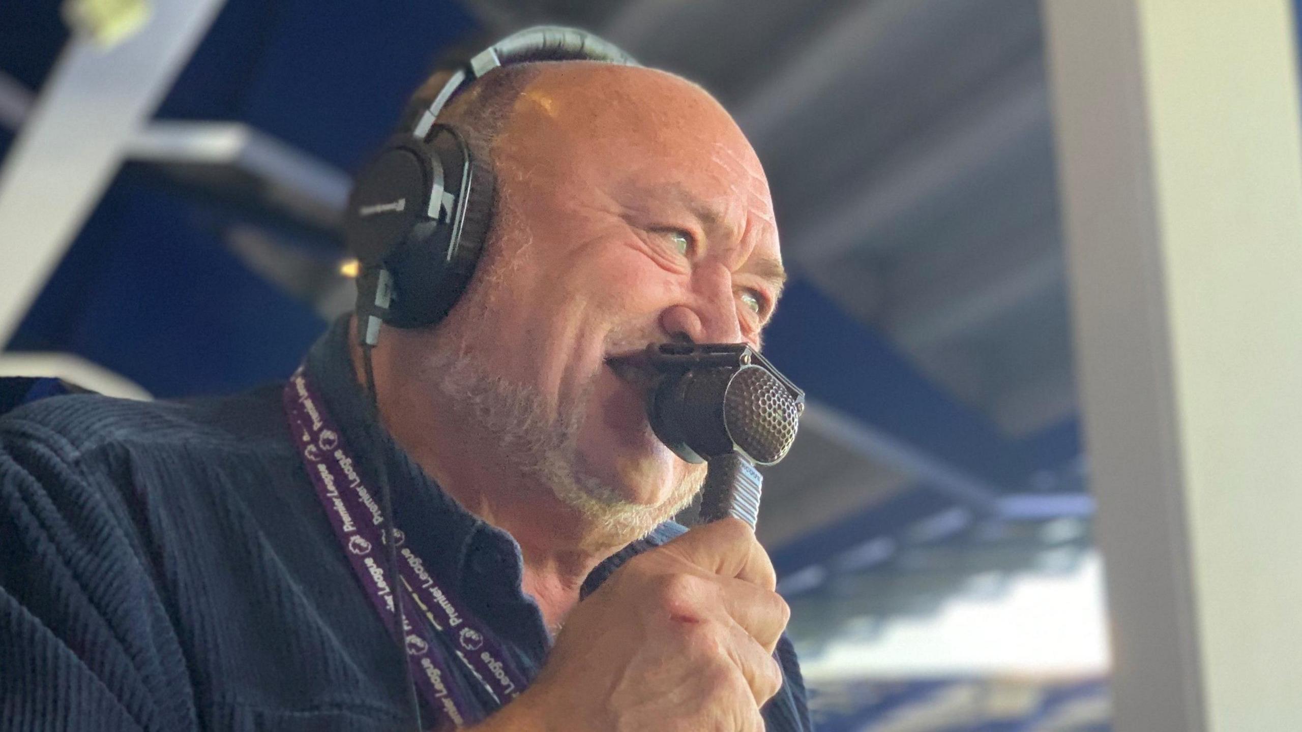 John Williams in the commentary box looking towards the pitch. He has a microphone pressed to his lips and is wearing headphones.