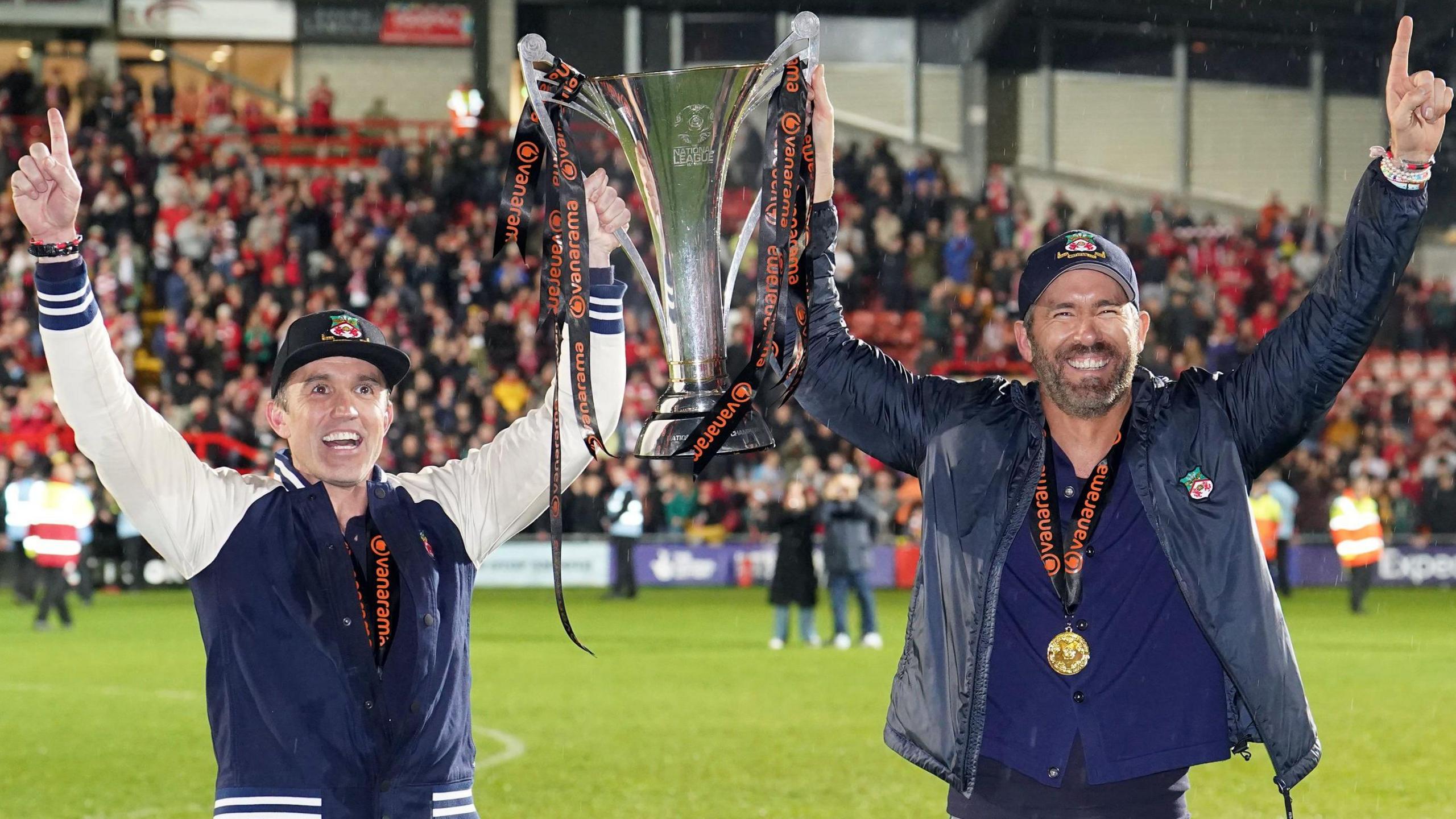 Ryan Reynolds and Rob McElhenney holding the National League Trophy on the pitch