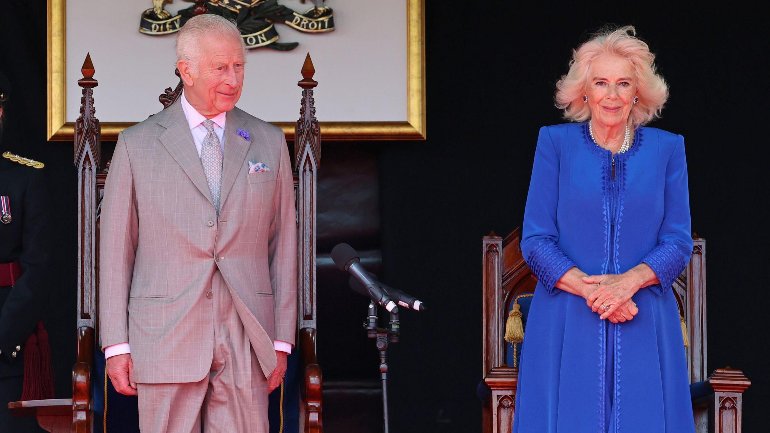 King Charles III and Queen Camilla pictured smiling at an event; the King wearing a grey suit and the Queen dressed in a bright blue coat.