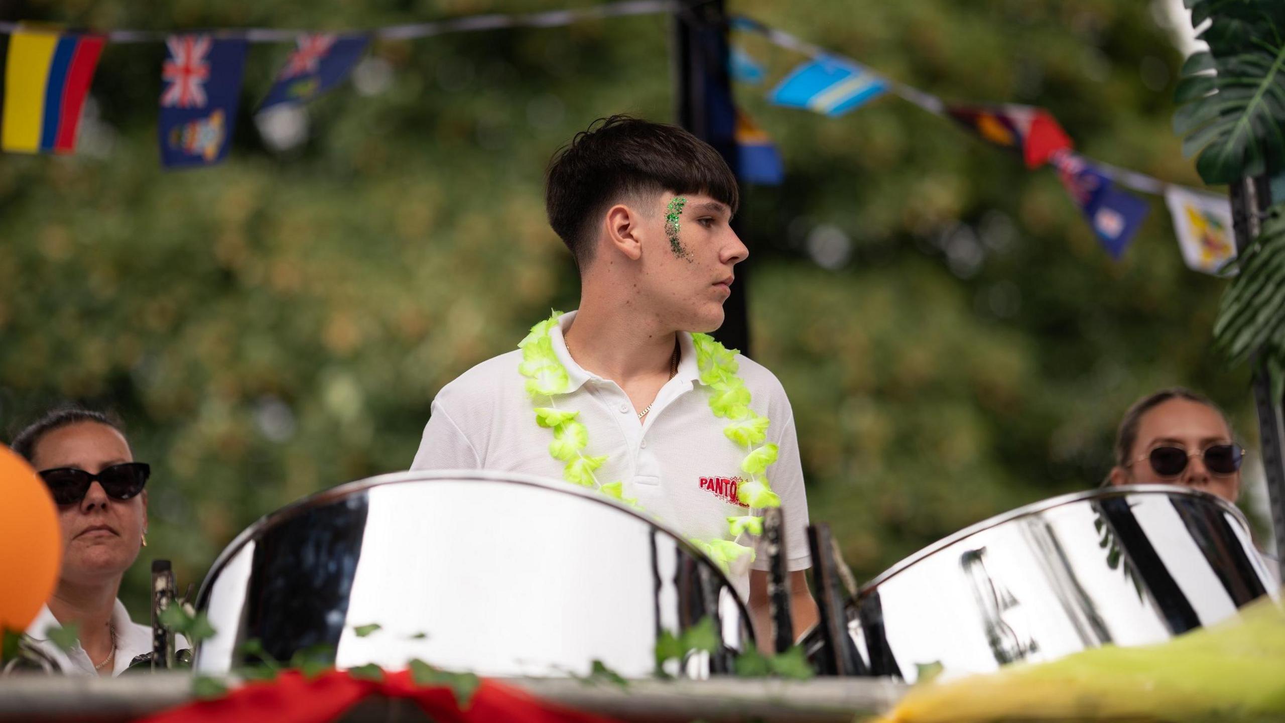 A young man wearing a white top and a yellow garland plays on two steel drums. He has a green glitter design on the side of his face and stands in front of bunting of various Caribbean flags fluttering overhead. Two women wearing sunglasses are behind with trees in the backdrop.