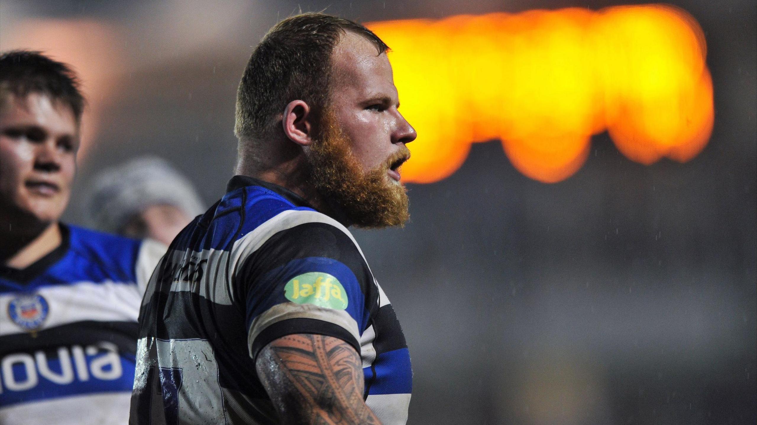 Jake Caulfield on the pitch at a Bath Rugby game. He wears a short-sleeved rugby shirt with the colours white, blue and black. He has red hair and a red beard and has a tattoo sleeve on his arm