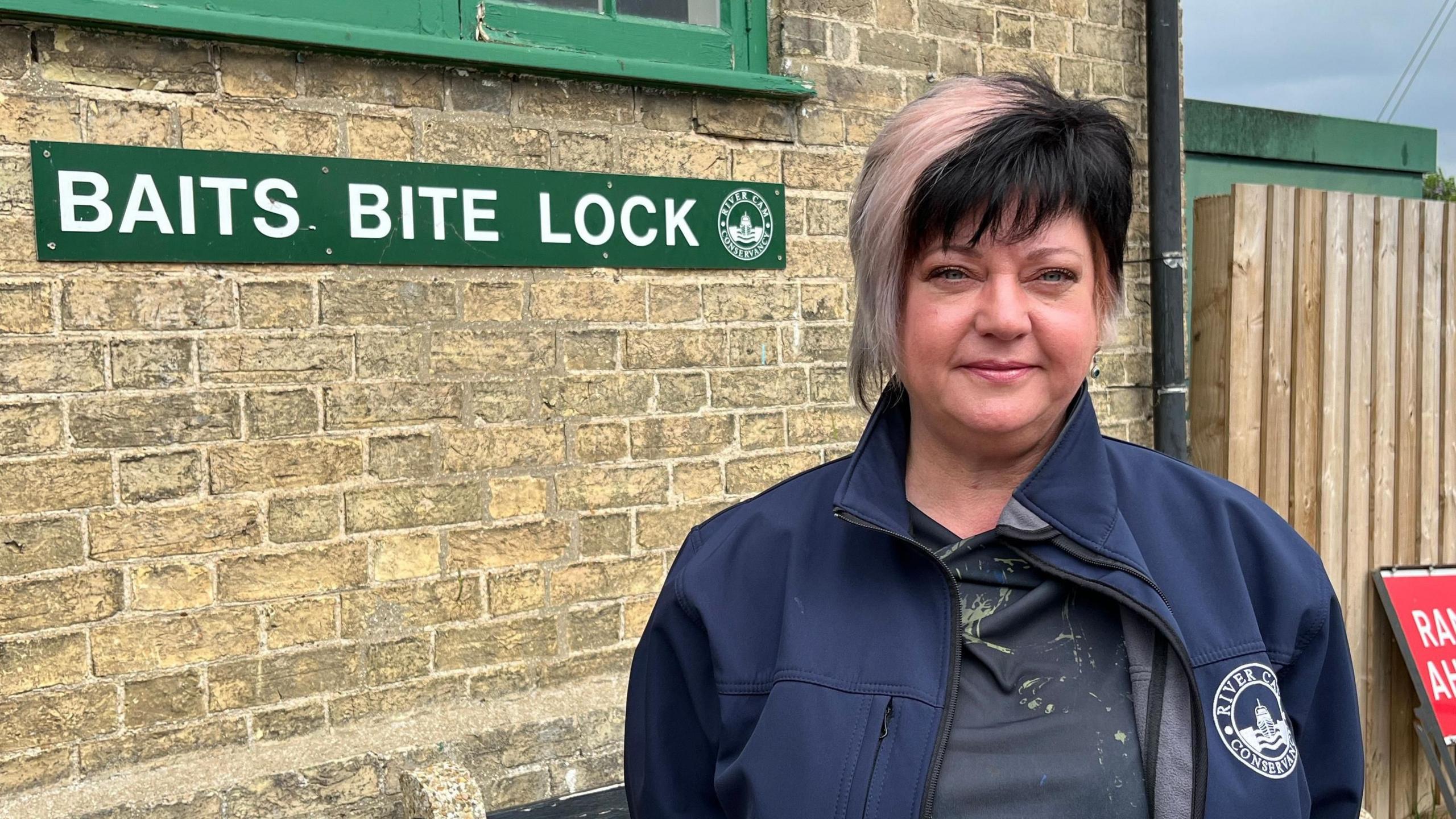 Helen has short black and white hair with hints of pink. She is wearing earrings and a blue jacket with the Conservators of the River Cam branding on the breast pocket. She is standing in front of a brick building and a fence, On the wall is a green sign reading Baits Bite Lock.