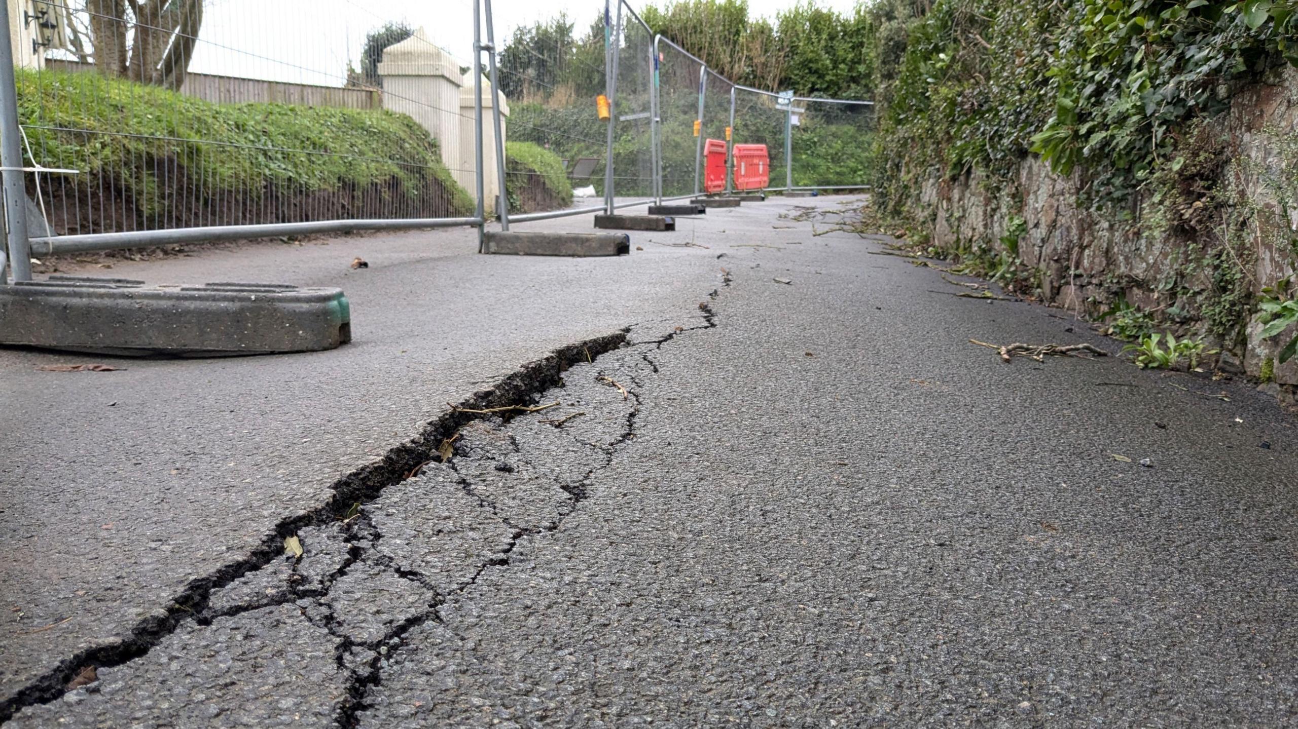 Various cracks run down the middle of a road as fencing and barriers blocks the road.