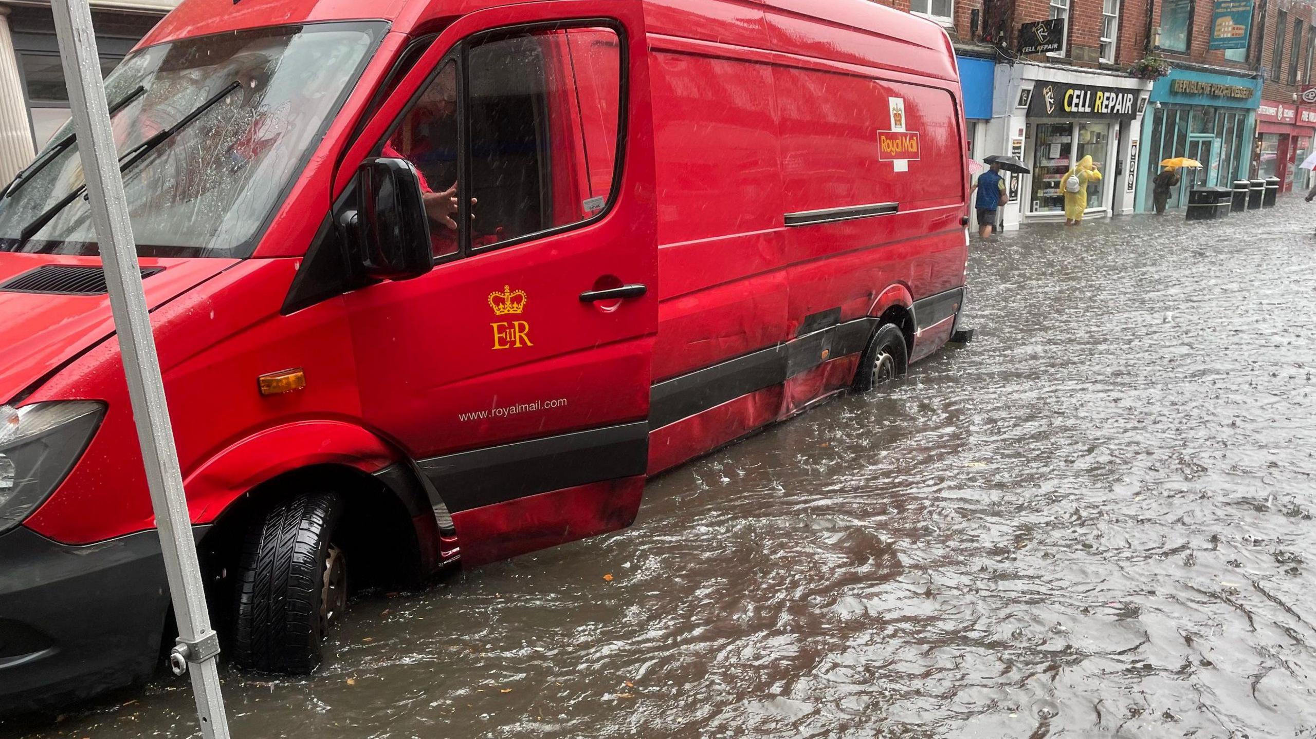 A red Royal Mail van abandoned in the high street with flooding water up to the wheels