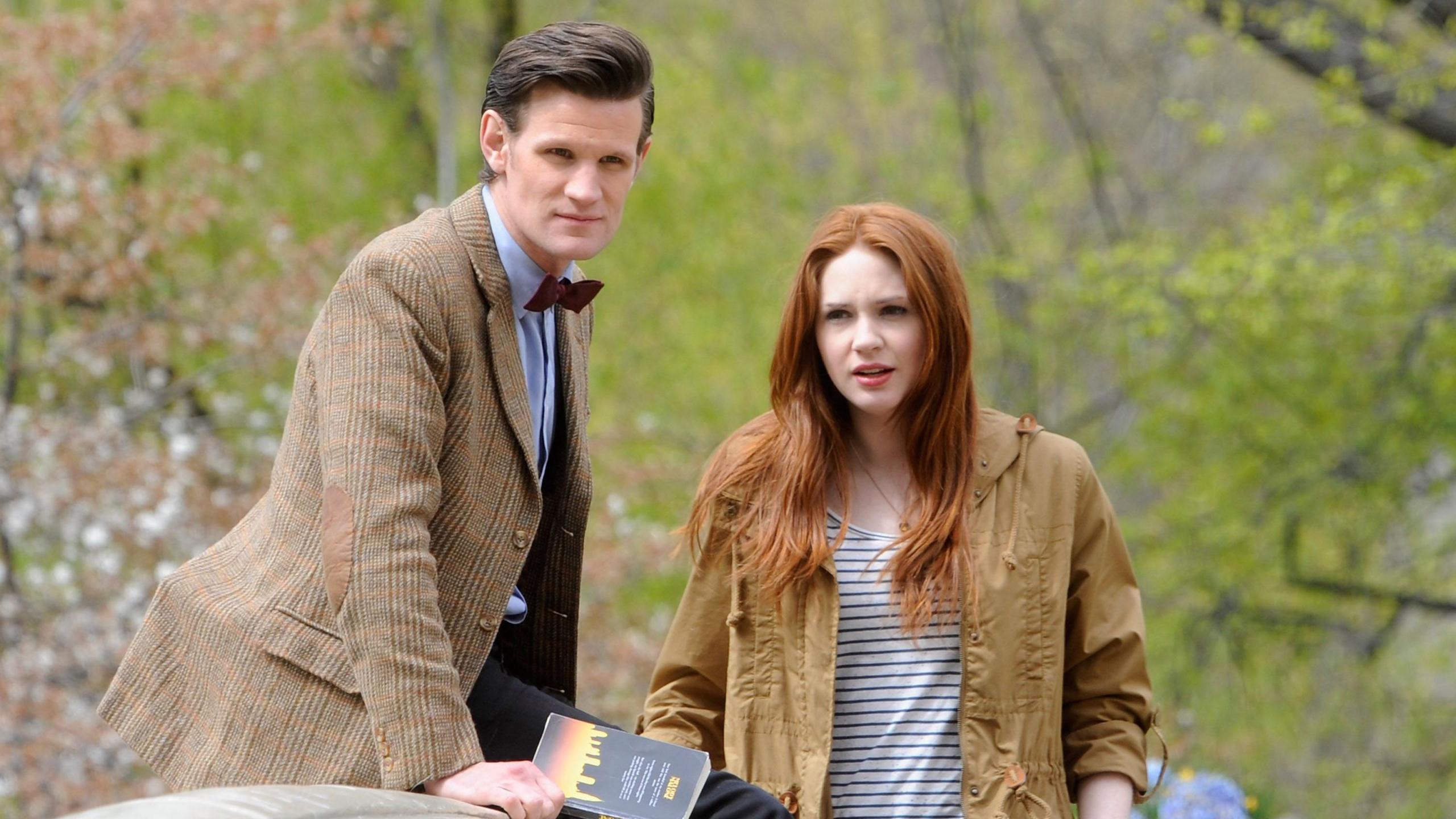 Matt Smith and Karen Gillan filming on location for "Doctor Who" in Central Park on April 11, 2012 in New York City. Matt wears a blue shirt with a dark red bow tie and a checked brown jacket. Karen wears a blue and white striped top and a brown anorak. They are outside in what appears to be a park.