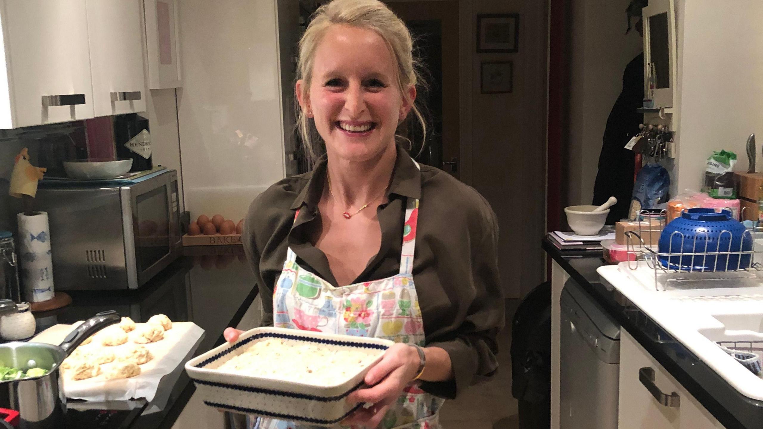 Lizzie Marsh standing in her kitchen wearing a brown shirt and apron, holding a ceramic dish and smiling at the camera