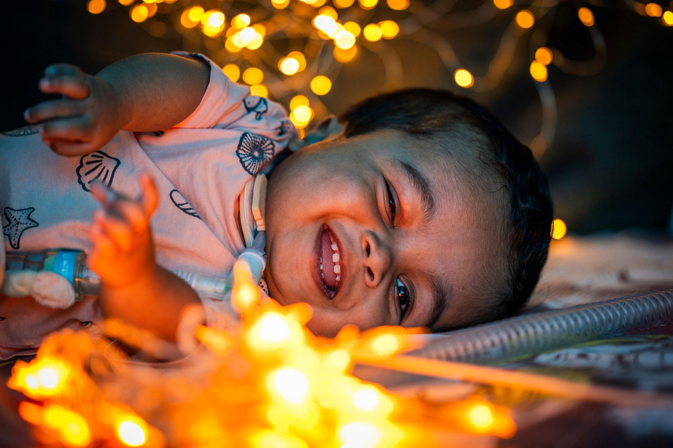 Gabriel is lying on a bed on his side. He is laughing and looking at lights that have been placed around his bed.