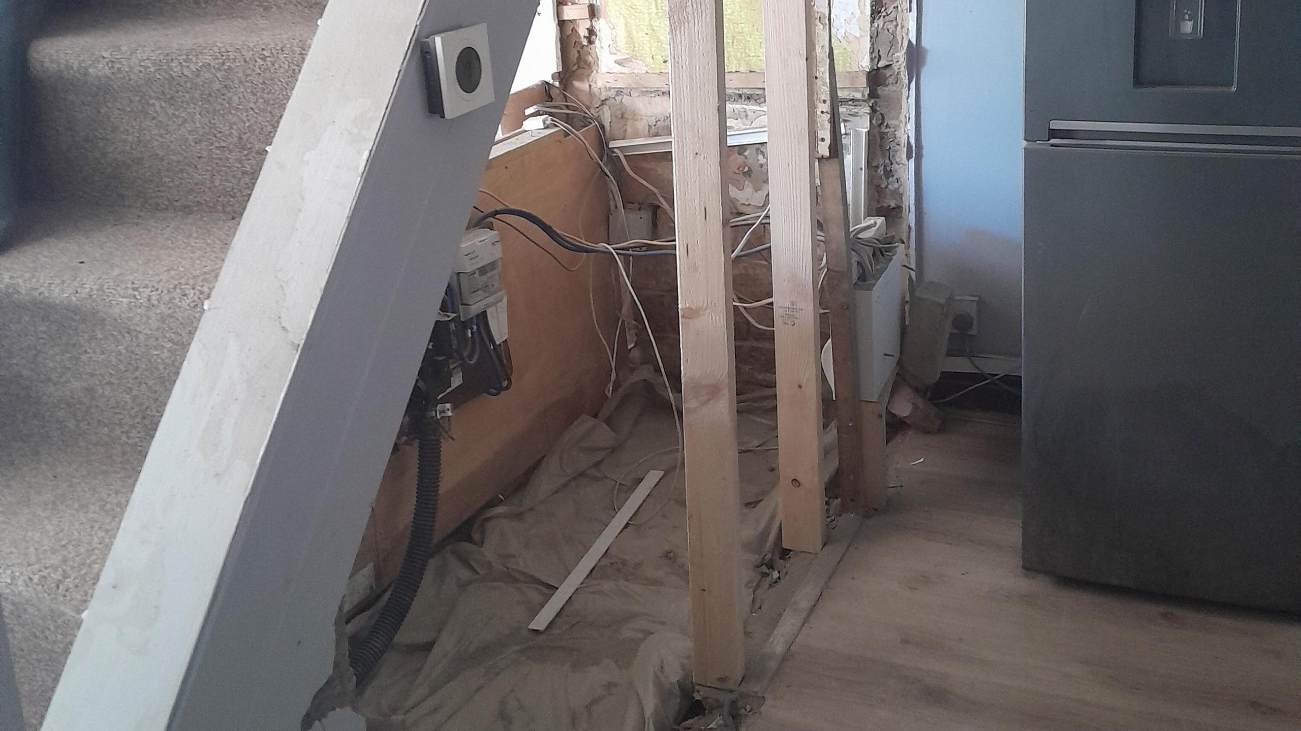 A set of stairs slightly to the left, but main focus is underneath them. The walls that would once have had an under stairs cupboard are gone - wooden poles are in place on one side, the fuse box attached to them with wires coming out the top and stretching across to a consumer board which is on a wooden board acting as a wall going halfway up.