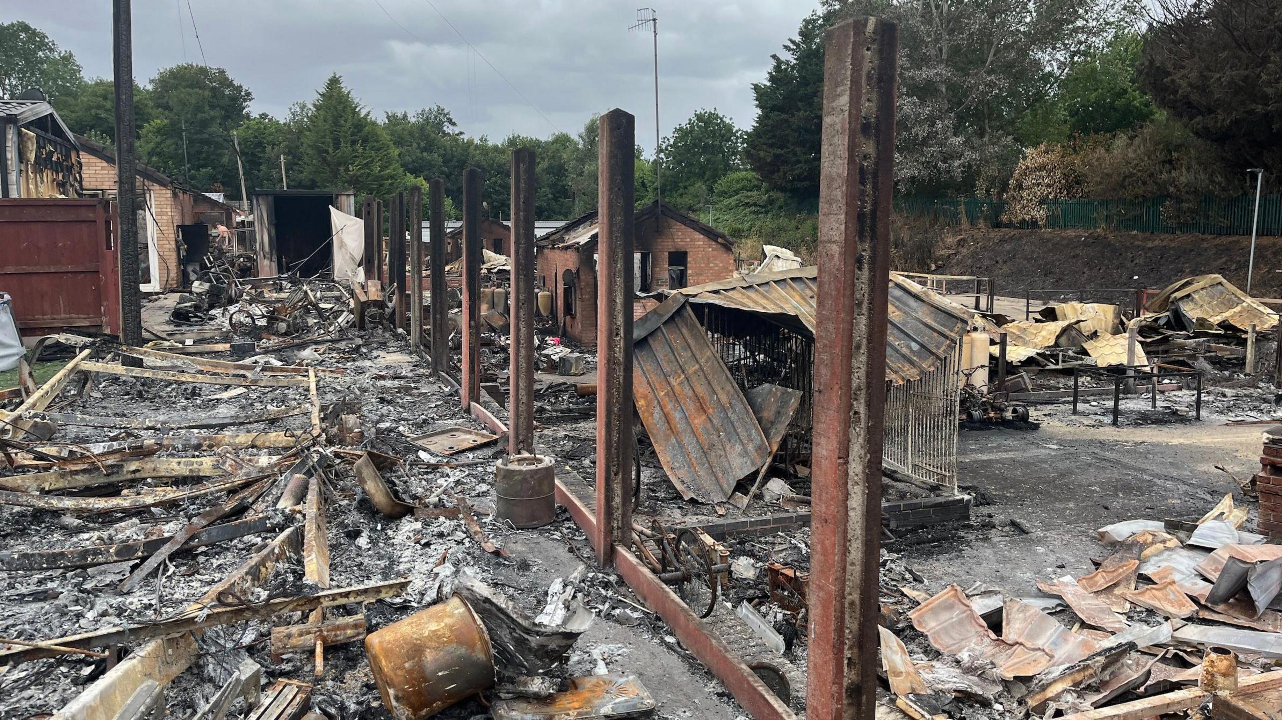 Fencing posts remain upright in the middle of the site, where the wooden panels were destroyed by fire. There is charred and blackened debris everywhere and trees in the background.
