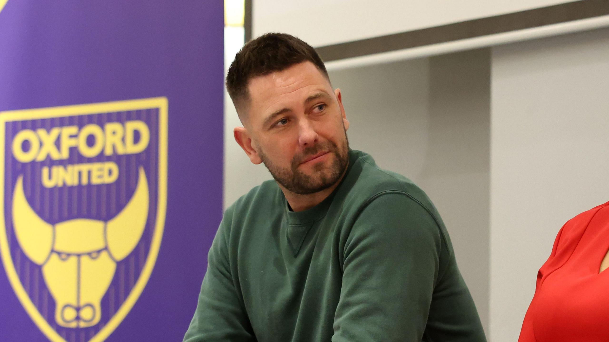 Des Buckingham wearing a green sweatshirt. He has short brown hair and stubble. Behind him is a yellow Oxford United logo.