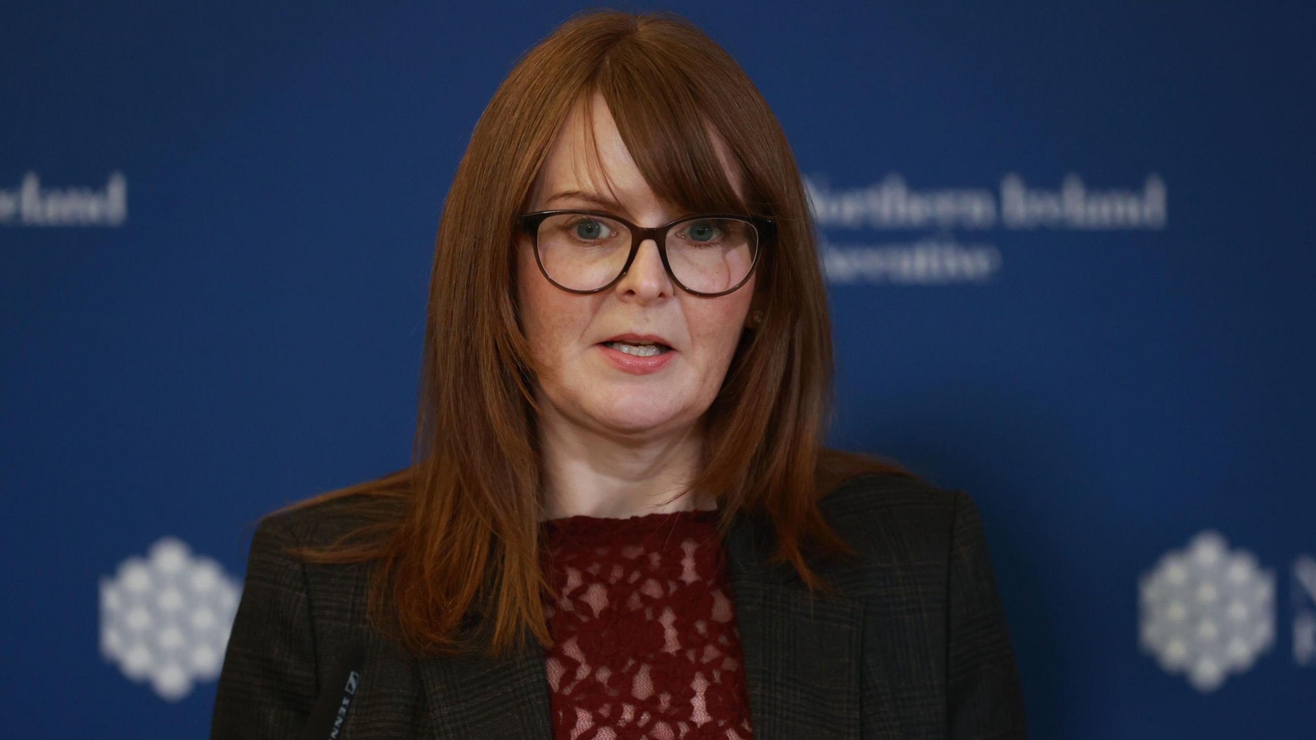 Caoimhe Archibald speaking at a microphone. She is wearing a grey blazer and red top and standing in front of a background with the logo of the Northern Ireland Executive