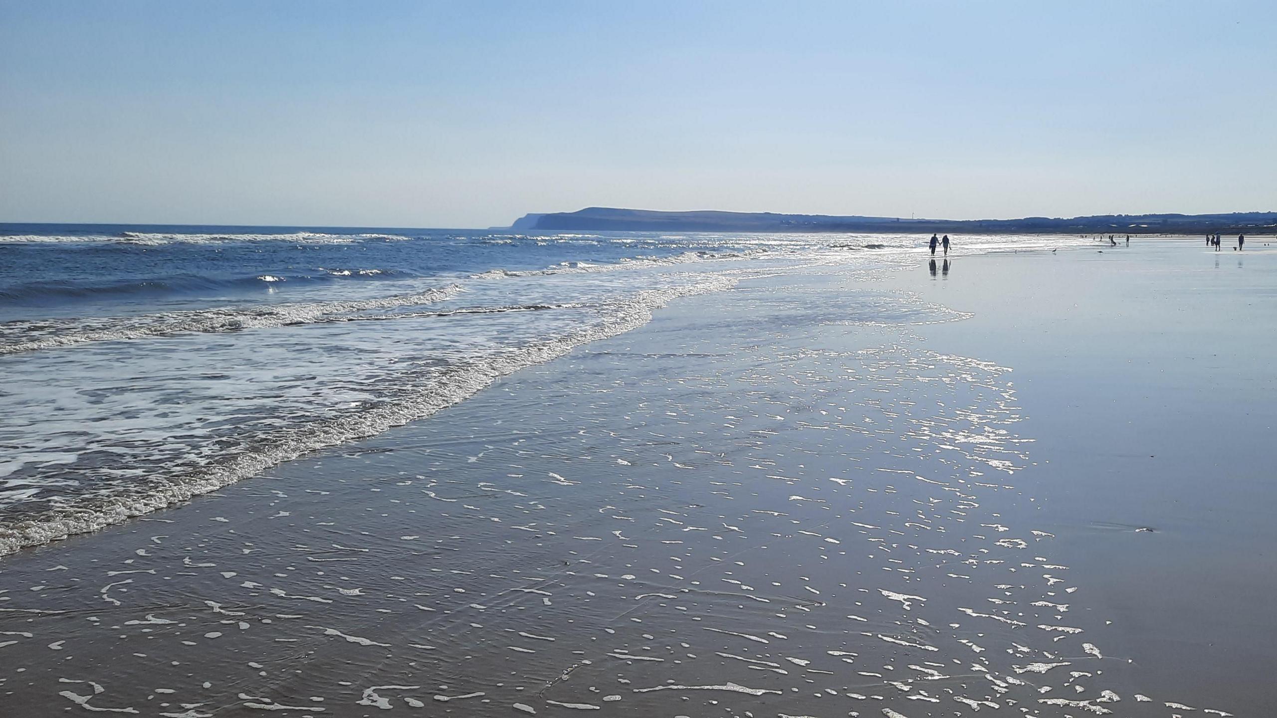 Photo of waves coming in at Marske-by-the-Sea with a blue sky above and people walking along the beach in the foreground